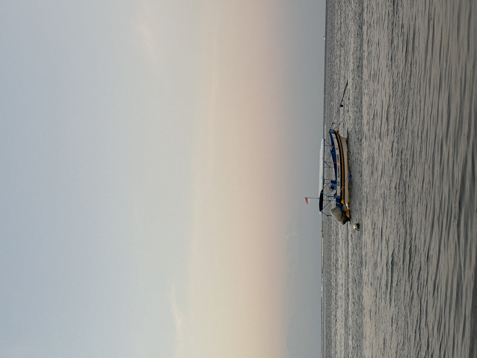 The image shows a long, narrow boat with a blue and white hull floating on a calm body of water. A small red and white flag is visible on the boat. The boat appears to be moored or anchored, as indicated by a fishing rod extending from its side and a buoy in the water nearby. The water has a rippled texture, suggesting gentle movement. In the background, the sky transitions from a pale grey near the horizon to a lighter, almost white hue further up. The lighting indicates it is either early morning or late evening, likely during a period of soft, diffused light, consistent with sunrise or sunset. The overall mood is serene and tranquil. The image composition is predominantly split between the vast sky and the expanse of water, with the boat serving as a focal point in the lower right quadrant. Text is visible on the side of the boat, reading "KUO JALA MANCA GANDHI 02". The location context of Kuta Selatan, Indonesia, suggests a tropical coastal setting.