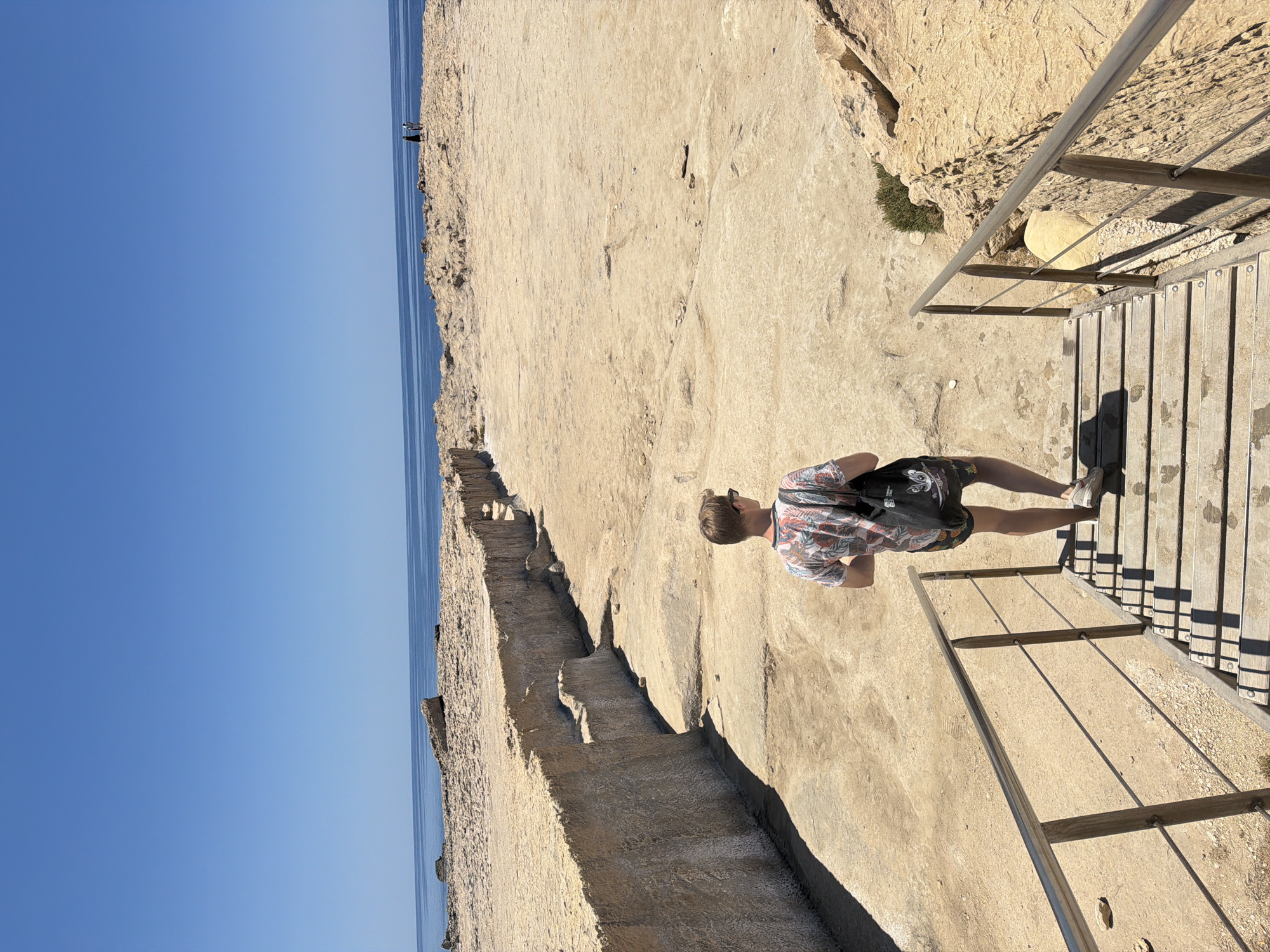 The media file shows a person standing on a wooden walkway with a metal railing. The person is wearing a short-sleeved, floral-patterned shirt and patterned shorts. They are also wearing white sneakers and a backpack. The walkway appears to be made of planks and runs alongside a rocky, sun-drenched cliff face. In the distance, the blue sea meets a clear blue sky.
The scene suggests a bright, sunny day, likely during daylight hours, given the strong shadows cast by the railing and the person. The rocky terrain and the presence of the sea are indicative of a coastal location. The walkway and railing suggest that this is a developed tourist area, perhaps a scenic viewpoint or a pathway along a cliffside.
In the far distance, near the edge of the cliff, a couple of small figures can be seen, possibly other visitors to the area. The overall impression is one of a relaxed outdoor scene, possibly a vacation or a day trip to a scenic spot. The lighting suggests it's a warm day. No visible text is present in the image. The location is specified as Saint Lawrence, Malta.