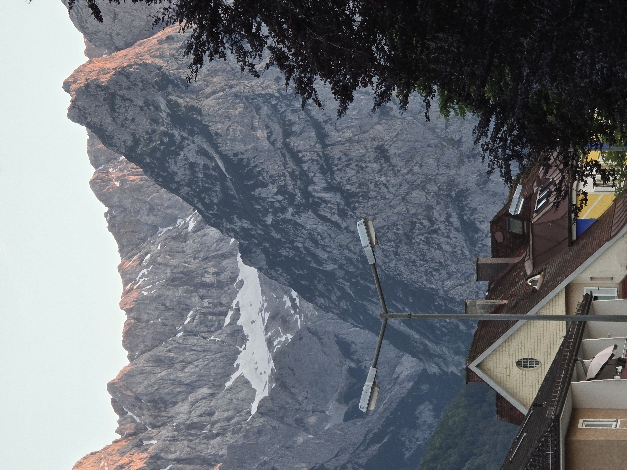 The image depicts a mountain range in Garmisch-Partenkirchen, Germany, bathed in the warm glow of sunrise. Jagged, rocky peaks, some still capped with snow, dominate the background. In the foreground, a street lamp stands prominently with its two lights extended. Below the lamp, the rooftops of several buildings are visible, featuring a mix of red tile and lighter-colored facades, including a house with an oval window. A dark, leafy tree occupies the right side of the frame, partially obscuring the mountains. The sky is a pale, clear blue, indicating early morning. There are no people or animals visible in the scene.