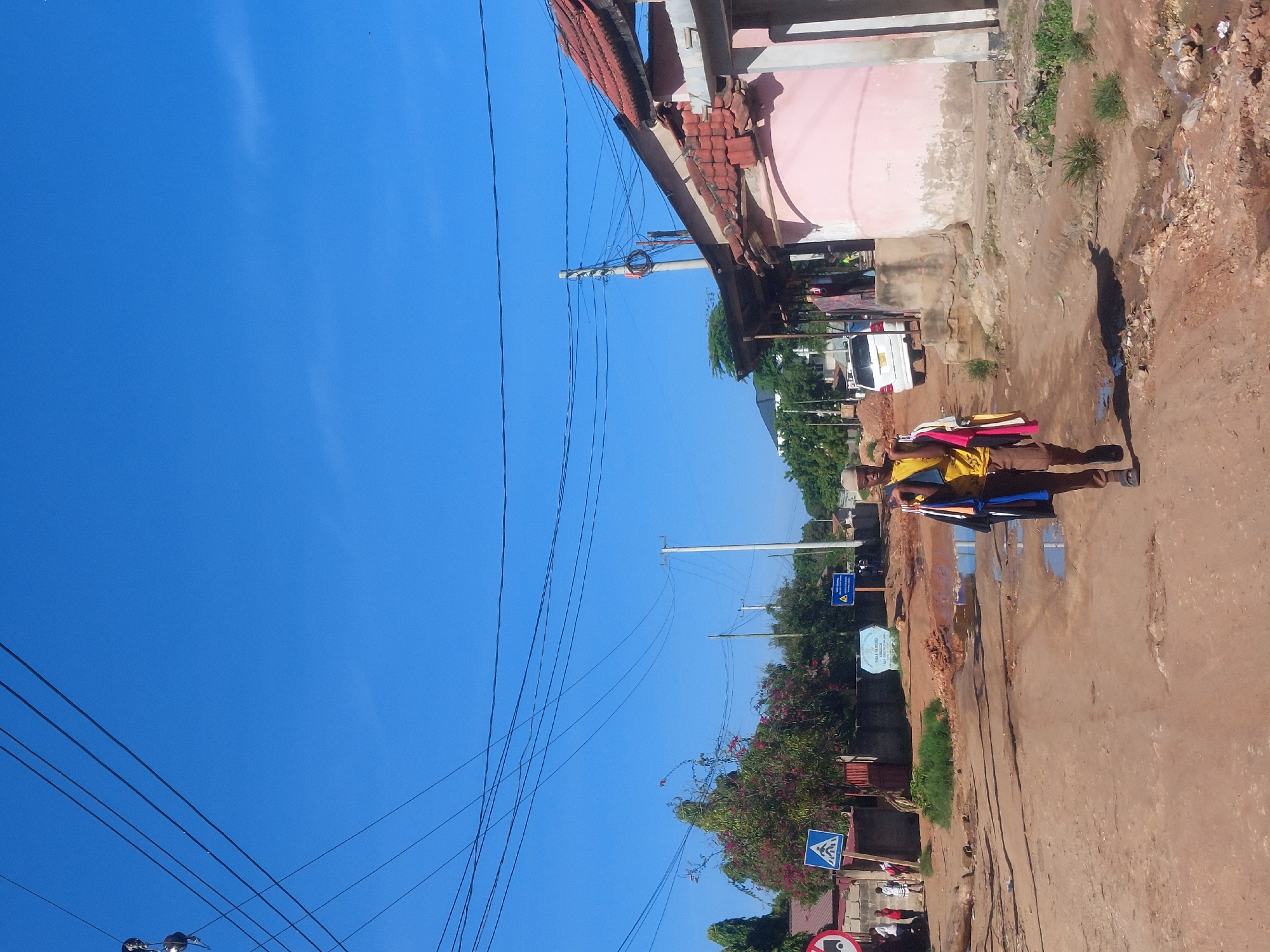 The image depicts a street scene in Dar es-Salaam, Tanzania. A man dressed in a yellow shirt, brown trousers, and a white cap walks along a muddy, red-dirt road. He carries several items, including what appears to be clothing or fabrics. The road shows signs of disrepair and puddles of water. Buildings line the street, one painted pink. Overhead are numerous electrical wires against a clear blue sky. Street signs are visible, including a pedestrian crossing sign. The overall environment appears to be a residential area.