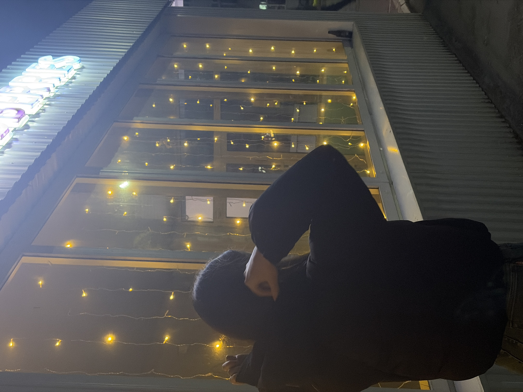 A low-angle, outdoor shot captures a person standing in front of a building at night. The building features a corrugated metal exterior with several large windows adorned with string lights. The person is wearing dark clothing and appears to be looking up at the window. A brightly lit sign is partially visible on the upper left corner. The scene appears to be at night, with ambient lighting creating reflections on the window surfaces. There is no indication of a location or context within Chaikovsky, Russia.
