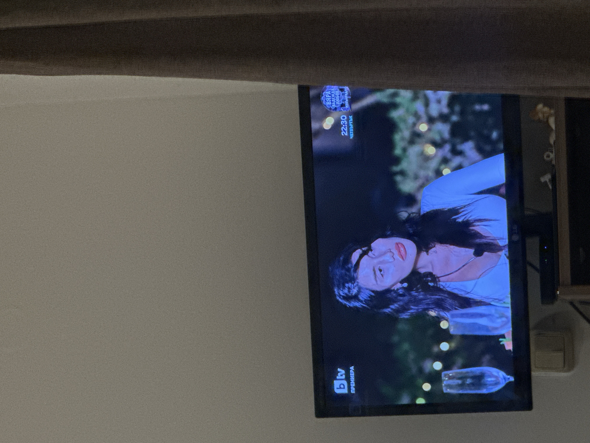 The media file presents an indoor scene featuring a television screen angled against a wall. A dark curtain or blind is visible across the top of the frame. To the right of the television, a section of a white wall and a light switch or power outlet are noticeable.
The television screen is displaying a program, prominently featuring a woman with dark hair and red lipstick, wearing a light-colored top. Her gaze is directed slightly upwards and towards the viewer. The background of the program content is dark with several blurred lights, hinting at an evening or night setting, possibly outdoors or in a decorative environment. Two glass-like objects, resembling bottles or decorative elements, are visible in the lower portion of the screen's image.
Several pieces of text are visible on the television screen, all in Bulgarian:
- In the top right corner of the screen: A circular logo with text including "BAPA AKADEMIYA" (VAPA ACADEMY), and smaller words like "БЪЛГАРИЯ" (BULGARIA), "ЛЮБОВ" (LOVE), "ИЗБОР" (CHOICE), "ИГРА" (GAME), "КЪСМЕТ" (LUCK). Below this, the time "22:30" and the day "ЧЕТВЪРТЪК" (THURSDAY) are displayed.
- In the bottom left corner of the screen: The logo for "b tv" (BTV), which is a Bulgarian television channel, and the word "ПРЕМИЕРА" (PREMIERE).
The overall impression is that of a TV program being watched in a home in Sofia, Bulgaria, likely late in the evening on a Thursday.