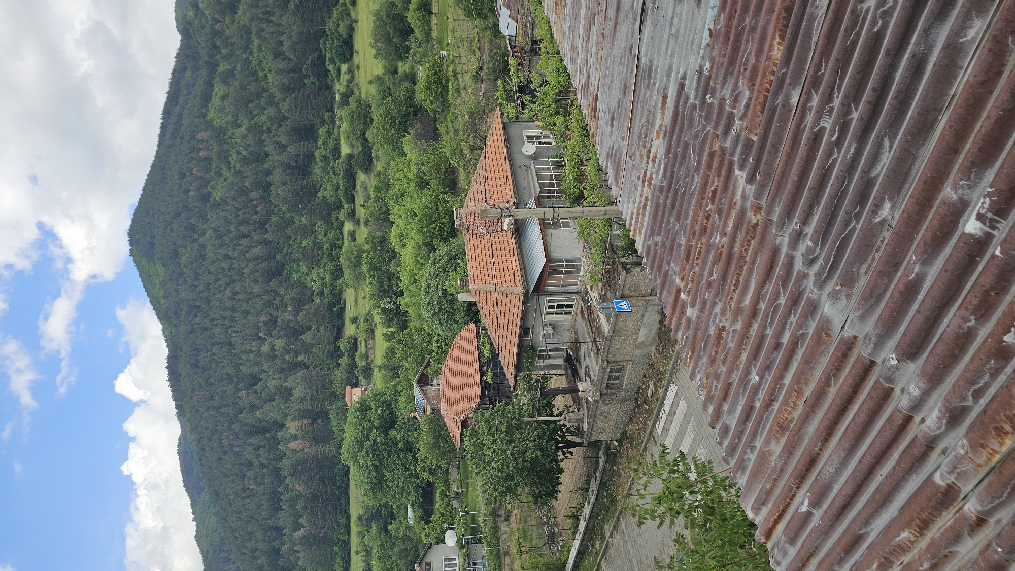 A high-angle, outdoor shot depicts a scene in Hristo Danovo, Bulgaria. The foreground is dominated by a corrugated metal roof, heavily rusted and textured. Buildings with orange tile roofs are nestled amidst dense green trees and vegetation in the midground, set against a backdrop of a steep, forested hillside rising to the left. The sky above the hillside is blue with scattered white clouds. A road cuts through the village near the buildings. A street sign stands in front of one of the buildings.
