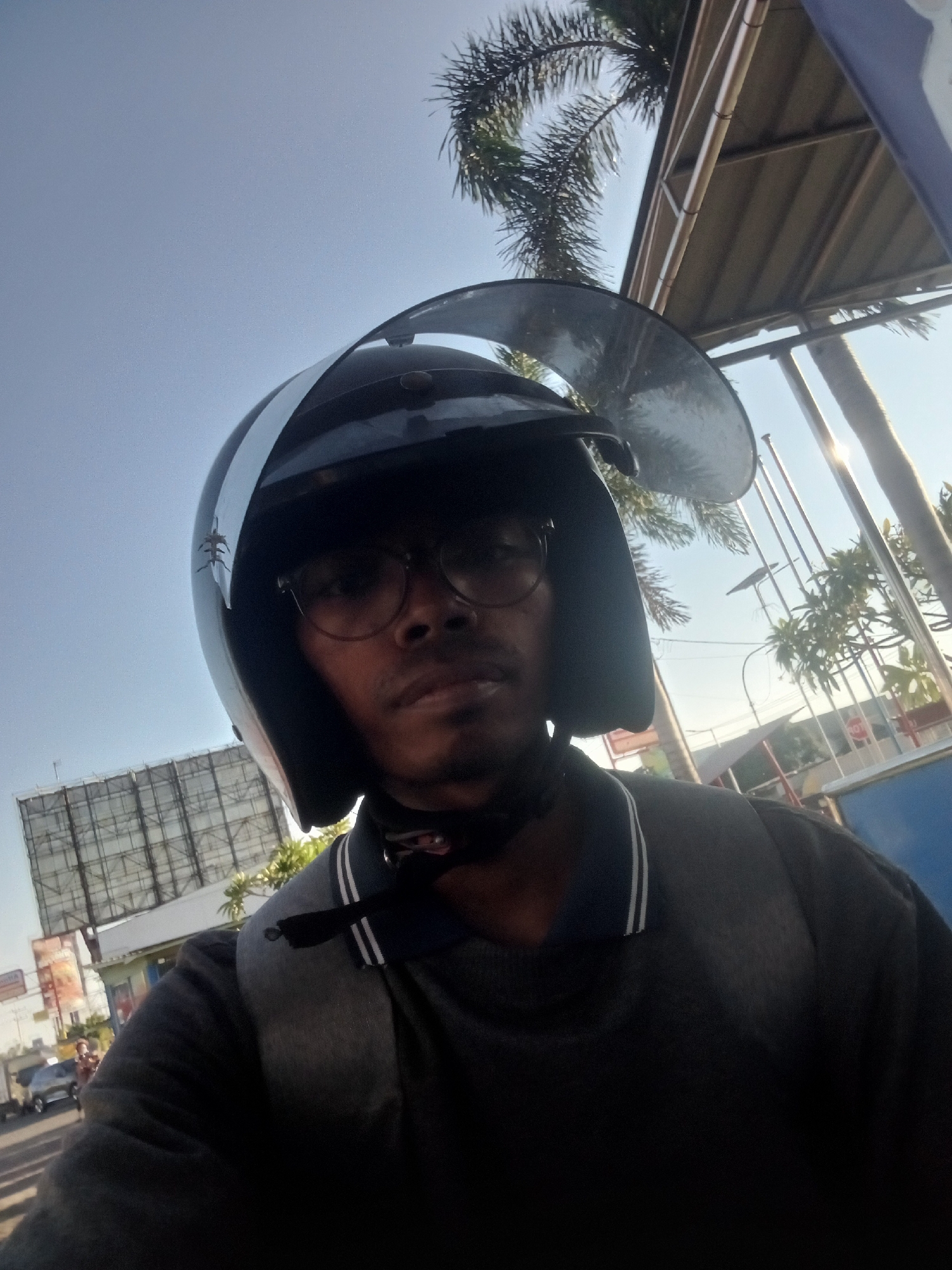 The image shows a selfie taken by a person wearing a black motorcycle helmet and glasses, with a gray sweatshirt. The person is looking directly at the camera. In the background, there is a large billboard on the left side of the frame, with a street and some buildings visible. Palm trees and a portion of a roof structure are also present. The sky is clear and blue, suggesting daytime. The scene appears to be an outdoor setting in a city, potentially on a street in Sawotratap, Indonesia, given the context provided. The person's facial features are partially obscured by the helmet and glasses.