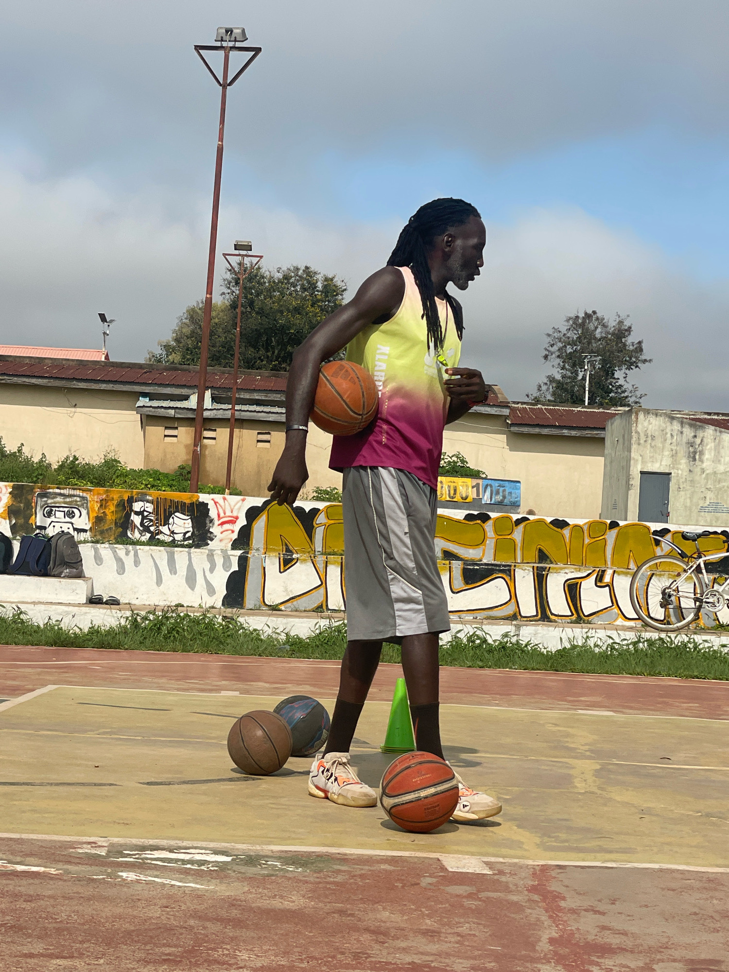 A tall, lean Black man with dreadlocks stands on an outdoor basketball court, holding a basketball in his left hand. He is wearing a yellow and pink sleeveless basketball jersey and grey athletic shorts. The court has a worn, yellow and red surface. There are three other basketballs on the court, one brown, one darker with a blue pattern, and another orange one near his feet. A green traffic cone is also visible near his feet. In the background, there is a white concrete wall covered in graffiti. Beyond the wall are some buildings with red roofs and trees. Two tall lampposts with lights are also visible against a cloudy sky. A bicycle is parked to the right of the graffiti wall. The scene appears to be an outdoor sports facility in a somewhat urban or semi-urban environment, possibly Gura Top, Nigeria, given the context.