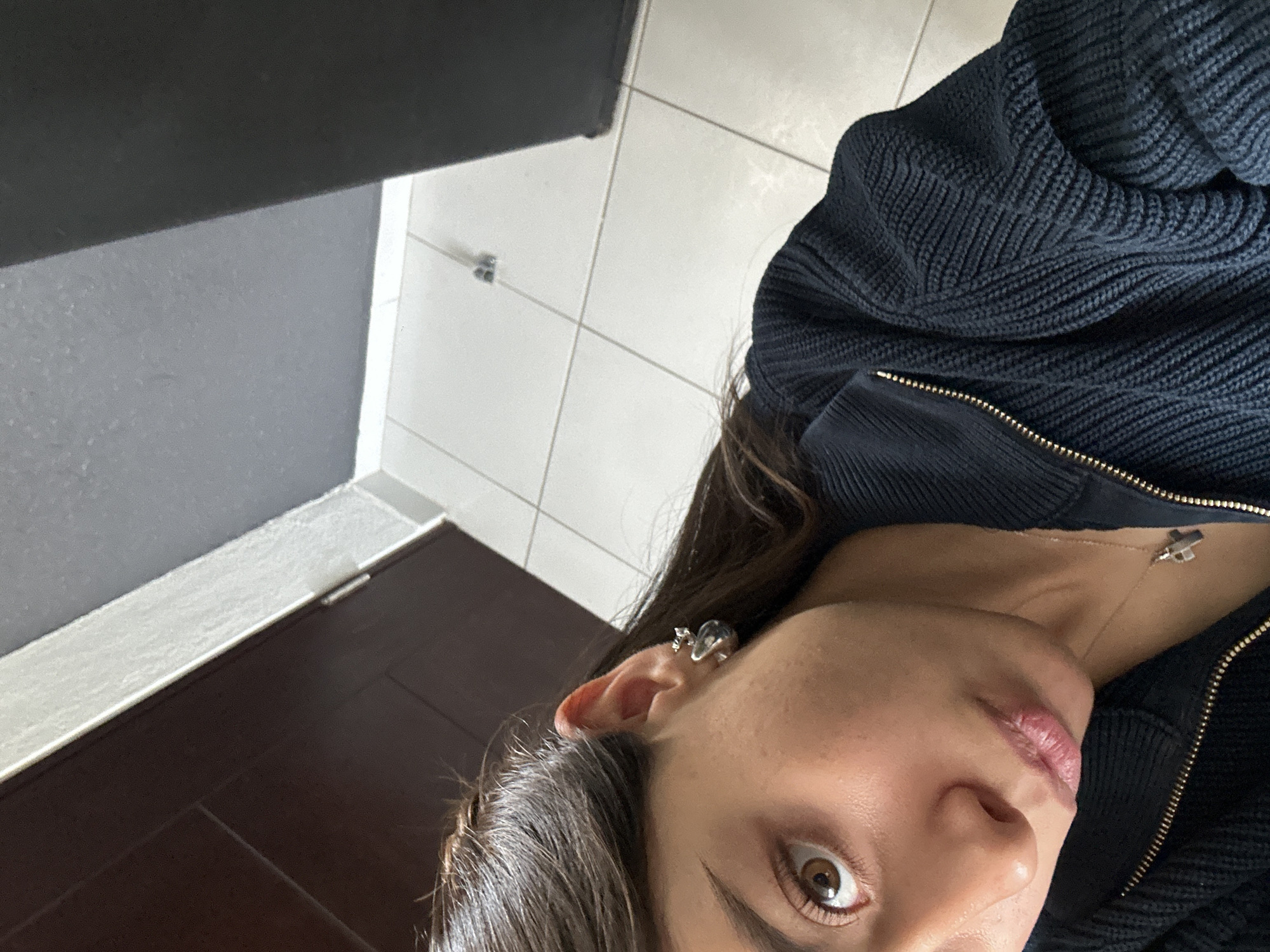A close-up, angled selfie of a person with fair skin, brown eyes, and dark brown hair wearing a dark blue ribbed jacket with a gold zipper. They are wearing an earring resembling a small animal in their left ear, and a necklace with a cross-shaped pendant. The background consists of a tiled floor, a dark gray wall, and a dark brown door. The location is likely indoors, possibly a bathroom, in an unknown city.