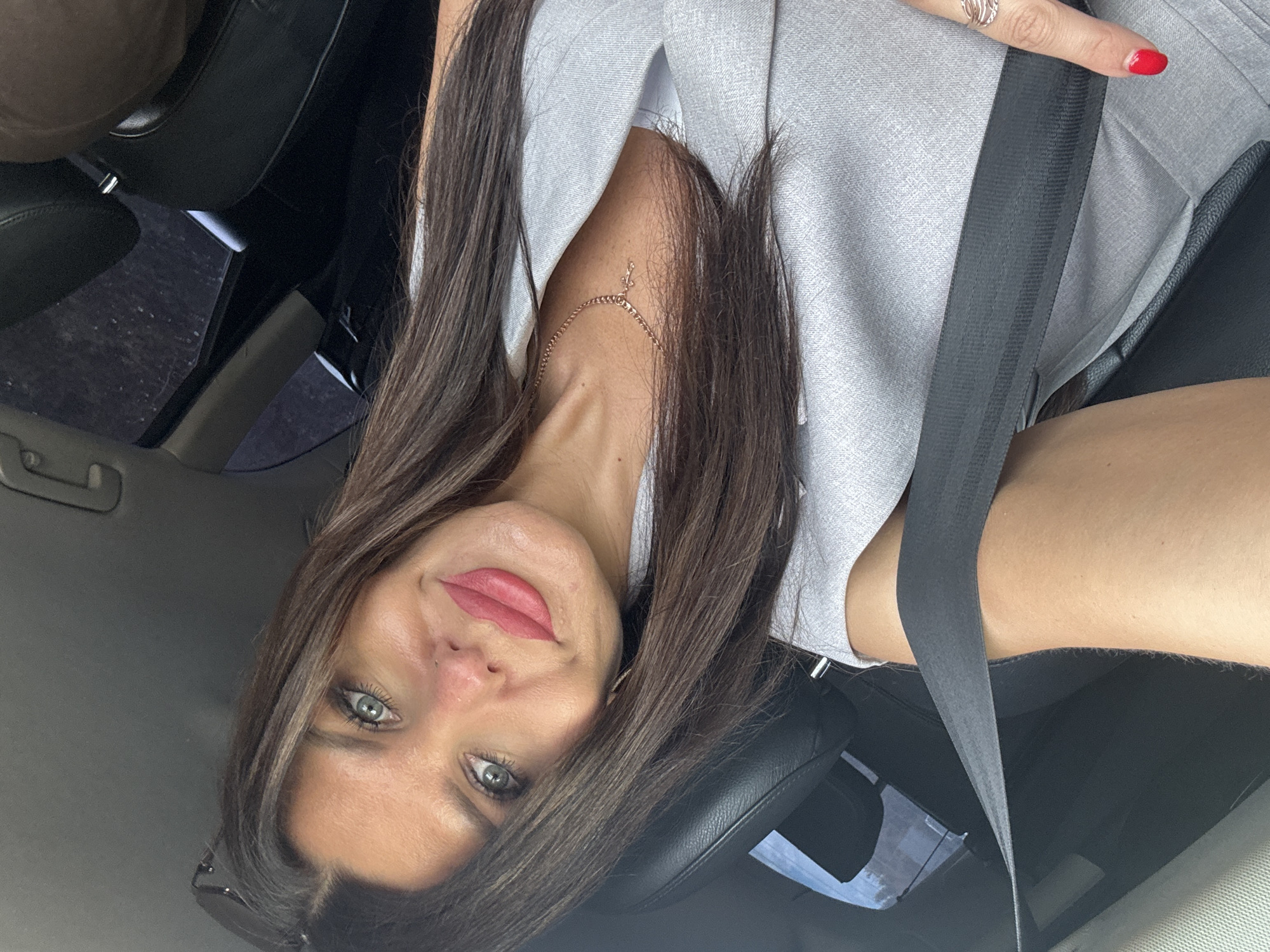 A woman is shown in a selfie taken from inside a car. She is wearing a light gray suit with a thin gold necklace. The seatbelt is across her body. Her makeup includes pink lipstick and blue eyeshadow. She has long, dark brown hair. The background suggests the interior of a vehicle, with black leather seats visible. The image does not contain information indicating a specific location in the city of Chaikovsky, Russia.