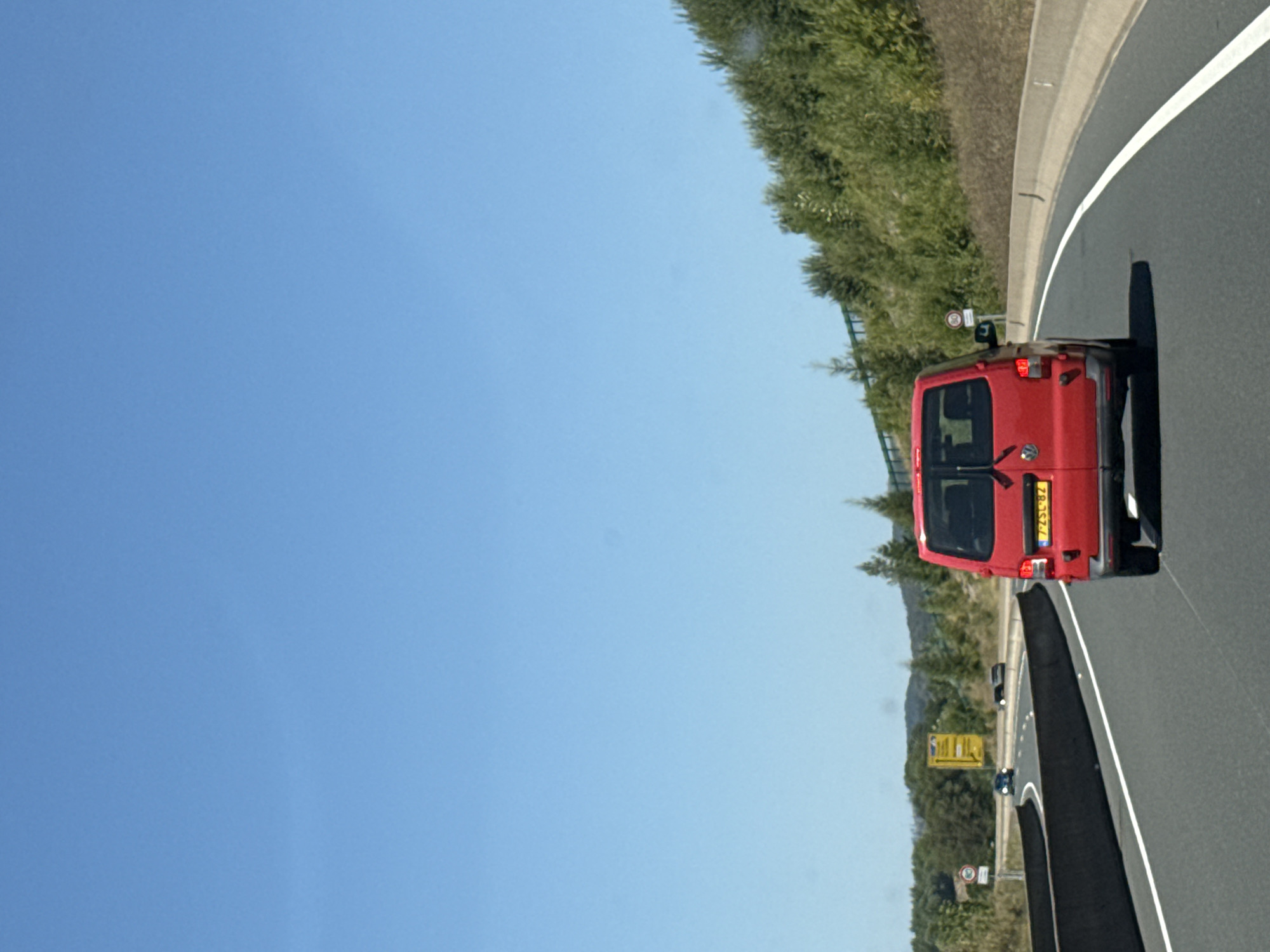 A red Volkswagen van is driving on a highway. The van has a yellow license plate with black lettering that reads "7-ZSL-82". The van is in the right lane of a multi-lane highway. The highway has a concrete barrier on the right side and a darker barrier on the left. The road is paved with asphalt and has white lane markings.
The background consists of a wooded hillside with green trees and bushes. The sky is clear and blue. There are signs visible along the side of the highway. One sign is a speed limit sign with a red circle and a white center with the number "80" in black. Another sign is yellow with black lettering. There are other vehicles on the highway in the distance.
The location is identified as the A7 highway near Bad Lauterberg im Harz, Germany. The scene depicts a typical highway driving environment under a clear sky.