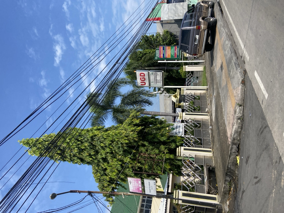The scene is a street corner in Kecamatan Tulangan, Indonesia (latitude: -7.481, longitude: 112.646). There is a single-story building with a green roof and a metal fence on the left side of the image. A large, leafy tree is located in front of the building. A palm tree is located behind the tree and building. There are many power lines running across the sky. A dark brown SUV is parked in the street. Several signs are visible including one sign that says "UGD" and another sign that says "PUSKESMAS KECAMATAN TULANGAN." The sky is blue with a few white clouds. The street is paved with asphalt. The sidewalk in front of the building is concrete.