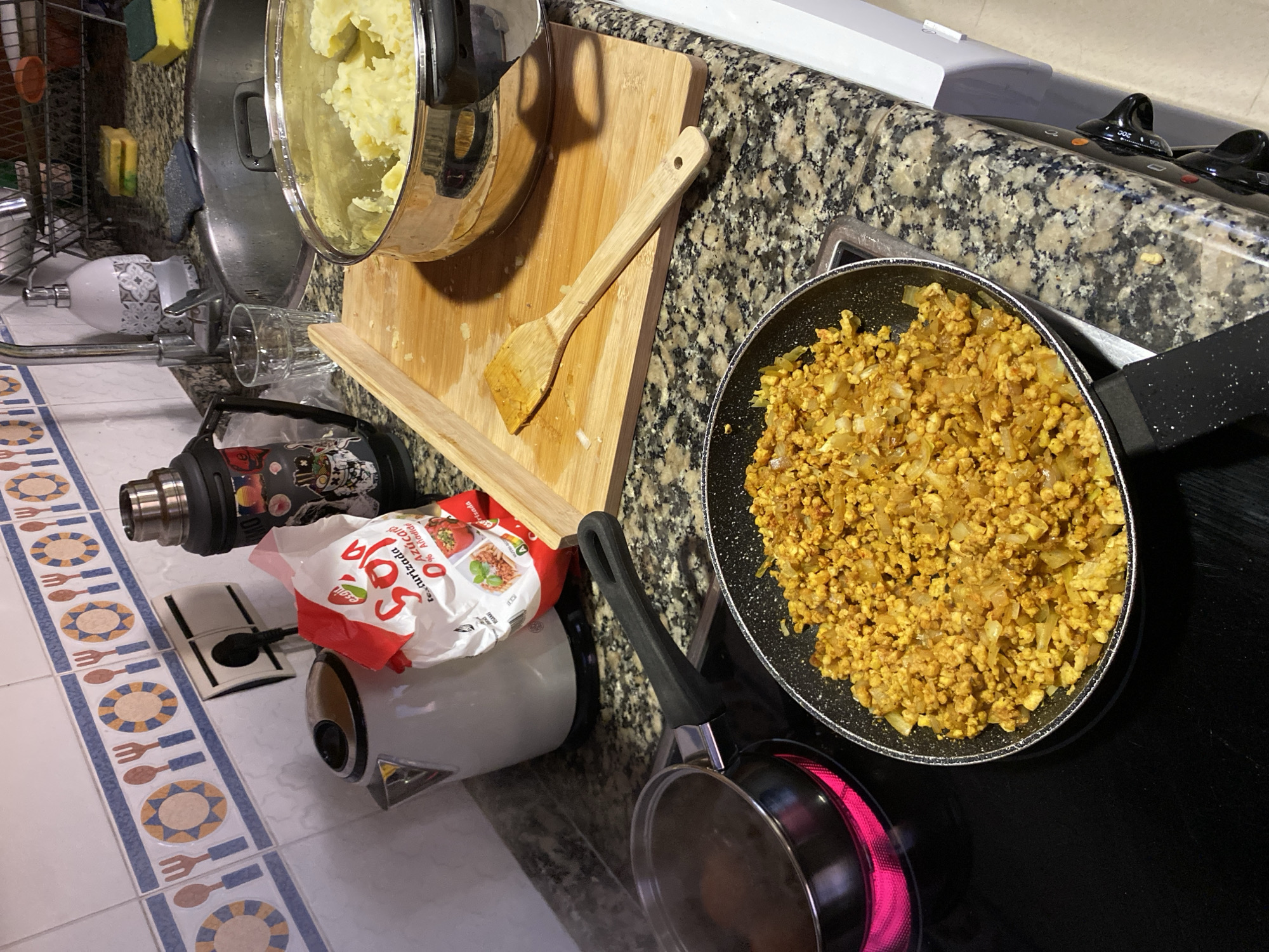 The image depicts a kitchen scene. A stainless steel pot containing mashed potatoes sits atop a wooden cutting board. A wooden spoon rests on the board as well. A package of soy granules is positioned near a white kettle. On the stovetop, a frying pan holds cooked food that appears to be a mixture of ground soy and onions. A smaller pot is visible beneath the frying pan. The stovetop is black, and the kitchen counter has a speckled gray pattern. The floor is tiled with a design of blue and orange circles and utensils. The image was likely taken indoors in an apartment. There is no basis in the image to infer the exact location in Gijón/Xixón, Spain.