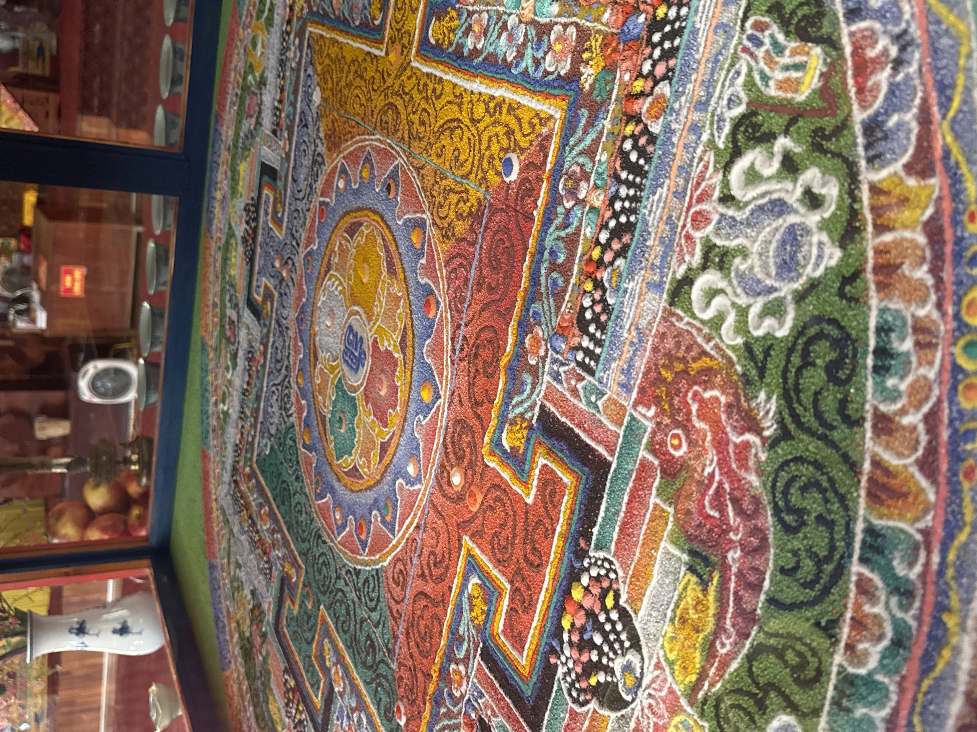 The image depicts a close-up, low-angle view of a sand mandala, a Tibetan Buddhist tradition. The mandala is intricately designed with vibrant colors and geometric patterns, crafted from finely colored sand. Its surface appears textured and granular. The mandala is enclosed within a clear glass casing, suggesting it is a display piece, possibly in a museum or temple. Reflections of the surrounding environment are visible on the glass, including a white vase with blue patterns, a stack of red apples, and a brass lamp. In the background, through the glass, there are hints of an indoor space with wooden furniture, a red sign, and a circular electronic device. The environment suggests a cultural or religious setting, likely a temple or a cultural exhibition space in Việt Trì, Vietnam, where sand mandalas are sometimes created and displayed.