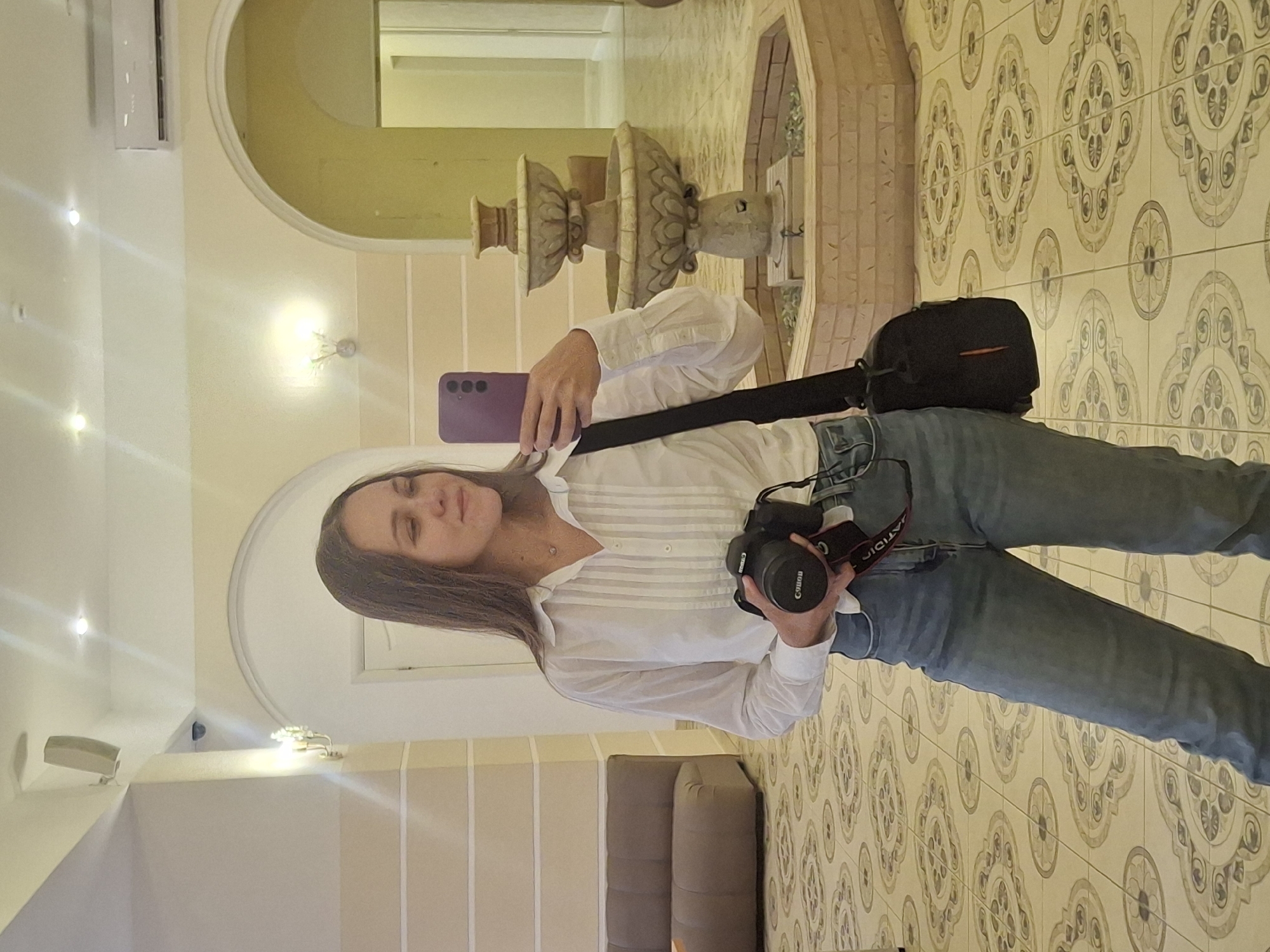 A young woman with long brown hair, wearing a white collared shirt and blue jeans, stands in an indoor atrium. She is holding a black Canon camera in her right hand and a purple smartphone in her left. A black camera bag is slung across her body. In the background, a multi-tiered stone fountain is visible, surrounded by a brick enclosure. The floor is tiled with an ornate pattern. The walls are painted in neutral tones with decorative sconces and arches. A portion of a brown sofa is visible on the left. The lighting appears to be artificial.
