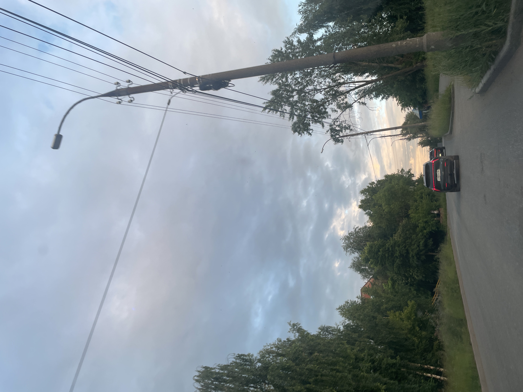 The image depicts an outdoor scene, likely in a semi-urban or rural area of Votkinsk, Russia. The perspective is from a moving vehicle, as suggested by the angle of the road and the blurred motion of the surroundings. The primary subjects are a road, lined with trees and tall grass, and a sky filled with dramatic, cloudy formations.
Prominent in the foreground and mid-ground are a series of utility poles and overhead wires, suggesting an electrical or communication network. One pole, leaning precariously, extends diagonally across the frame, its curved arm holding a streetlamp. The sky, a mix of light and dark clouds, indicates a possibly overcast or stormy weather condition, with hints of sunset colors near the horizon, suggesting it's either early morning or late evening.
Two dark-colored cars are visible on the road, one in the distance and another closer to the foreground, their brake lights illuminated. Dense green trees and foliage flank the road, with patches of visible grass. In the background, peeking through the trees, are glimpses of buildings, one with a red brick facade. The overall mood is somewhat subdued, possibly due to the weather and the time of day, with a sense of movement and journey. No discernible text is visible in the image.