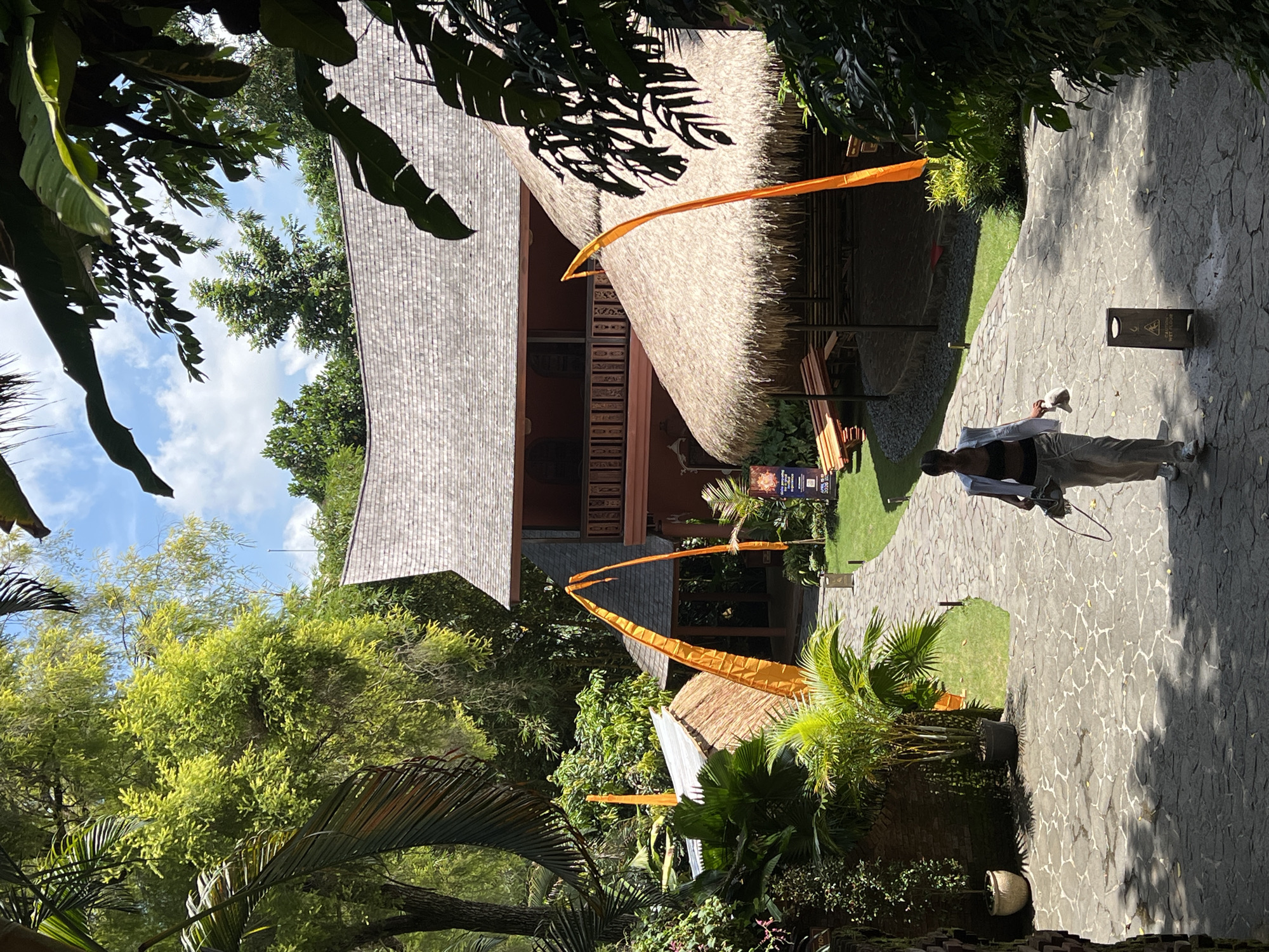 The photograph was taken in Peliatan, Indonesia. It features a stone path winding between tropical foliage and traditional Balinese buildings. One building has a grey tiled roof and exposed dark-wood beams. Another structure has a thatched roof and decorative orange flags attached to its peaks. A person in light clothing is walking along the path, holding a small object that may be a dog. A sign indicating caution is visible on the path.