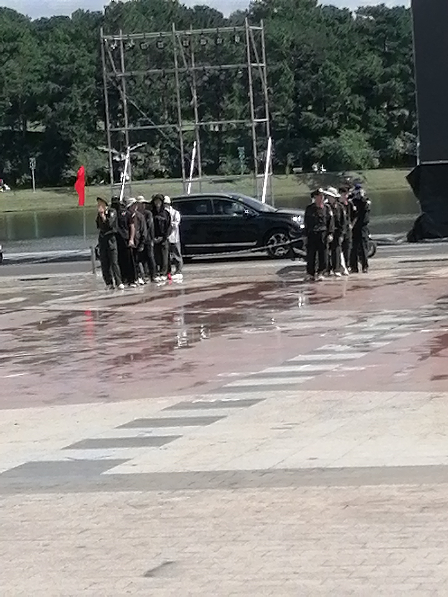 In Da Lat, Vietnam, a group of individuals, likely in uniform, stands in formation on a wet paved area near a black car. Behind them, a large metal scaffolding structure with lights is set up, and beyond that, a body of water and lush green trees form the backdrop. A red flag is visible on a pole to the left of the scaffolding. The ground is reflective, suggesting recent rain. The individuals are facing away from the viewer, and their uniforms appear to be dark.