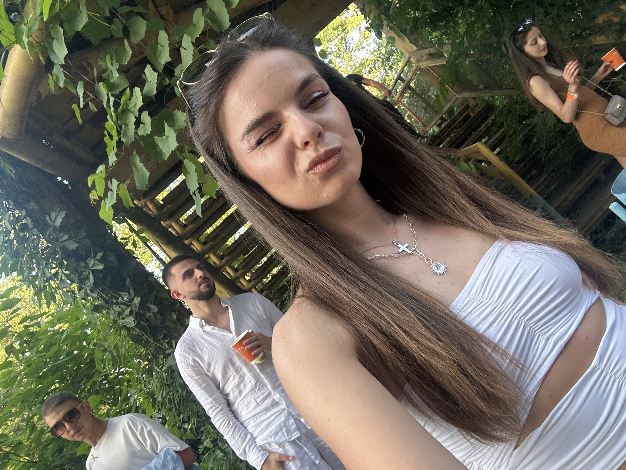 The photograph depicts a group of people outdoors in what appears to be a partially wooded area. In the foreground, a young woman with long brown hair, wearing a white top and silver necklace, is posing for a selfie or photo, with one eye closed and lips pursed. Behind her and to the left, two young men are visible. One is wearing a white shirt and holding a beverage cup, while the other wears sunglasses and a white t-shirt. In the background, to the right, a woman in a brown dress is seen holding a phone or small device. A wooden structure, possibly stairs or a viewing platform, is partially visible in the background, along with green foliage and trees.
Given the provided location of Plovdiv, Bulgaria, and the presence of outdoor structures and greenery, it's possible that the scene is situated in one of the city's parks or recreational areas. However, without further details or specific landmarks, pinpointing the exact location is challenging.