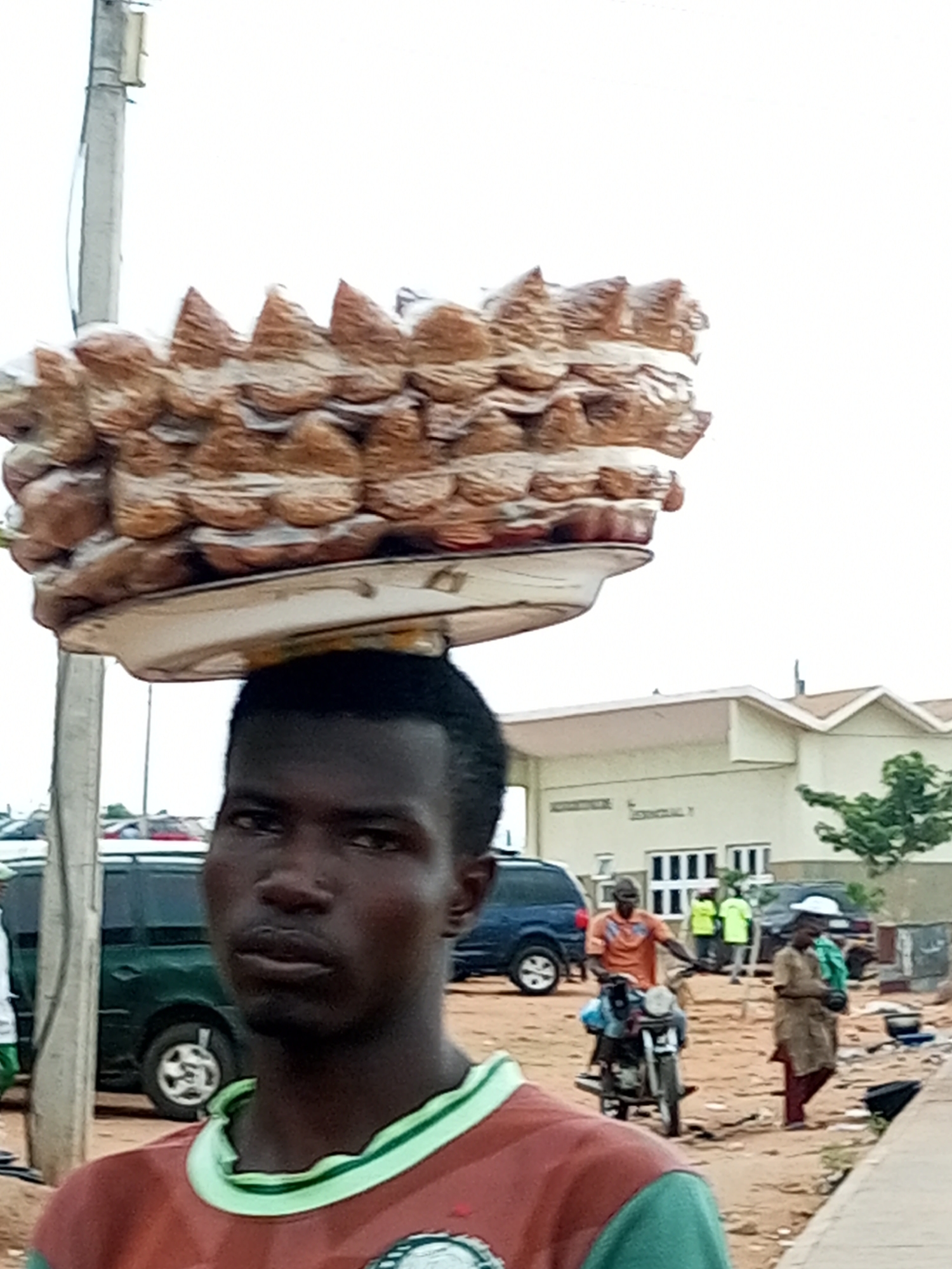 In Gombe, Nigeria, a man carries a large load of baked goods balanced on his head, a common sight in bustling markets. The goods, likely bread or a similar baked item, are stacked high and wrapped in plastic. He is the central focus, with his face partially visible. In the background, a dusty street scene unfolds. A green minivan is parked, and other vehicles are visible further down the road. Several people are in the distance, including a man on a motorbike, another in a bright green shirt, and a woman in a long garment. A building with a light-colored facade and a brown roof stands in the background. The overall environment suggests an outdoor market or a busy street in a Nigerian city.