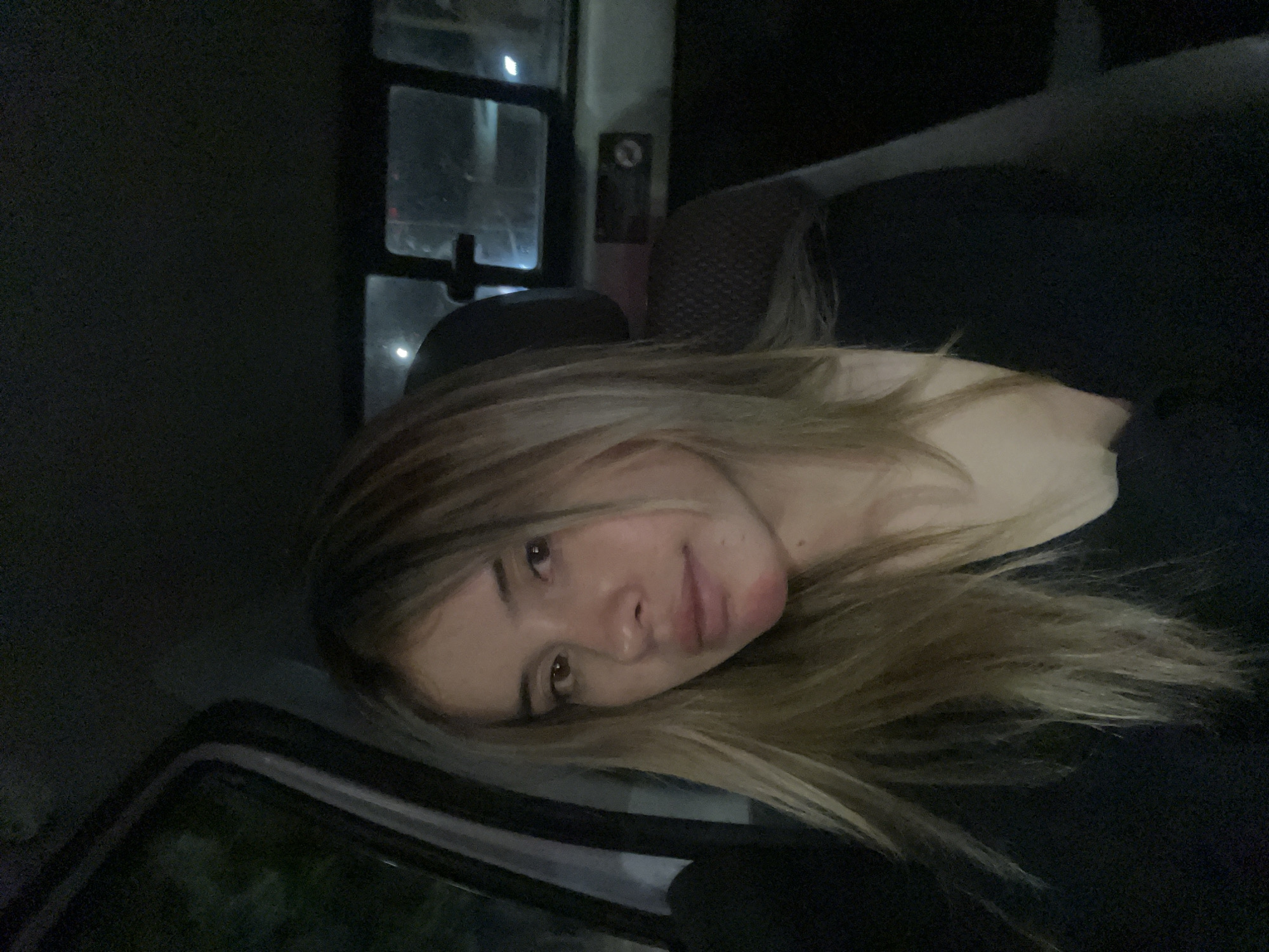 A young woman is in the front seat of a vehicle. She is leaning her face on the edge of the open window, facing toward the camera with a slight smile. The location is likely in San Fernando del Valle de Catamarca, Argentina, although there is no physical evidence within the photograph that confirms this. It is dark outside. The interior of the vehicle is dark, with the details visible in the interior of the cabin. Part of the vehicle's seat is visible. There is a window behind the seat.