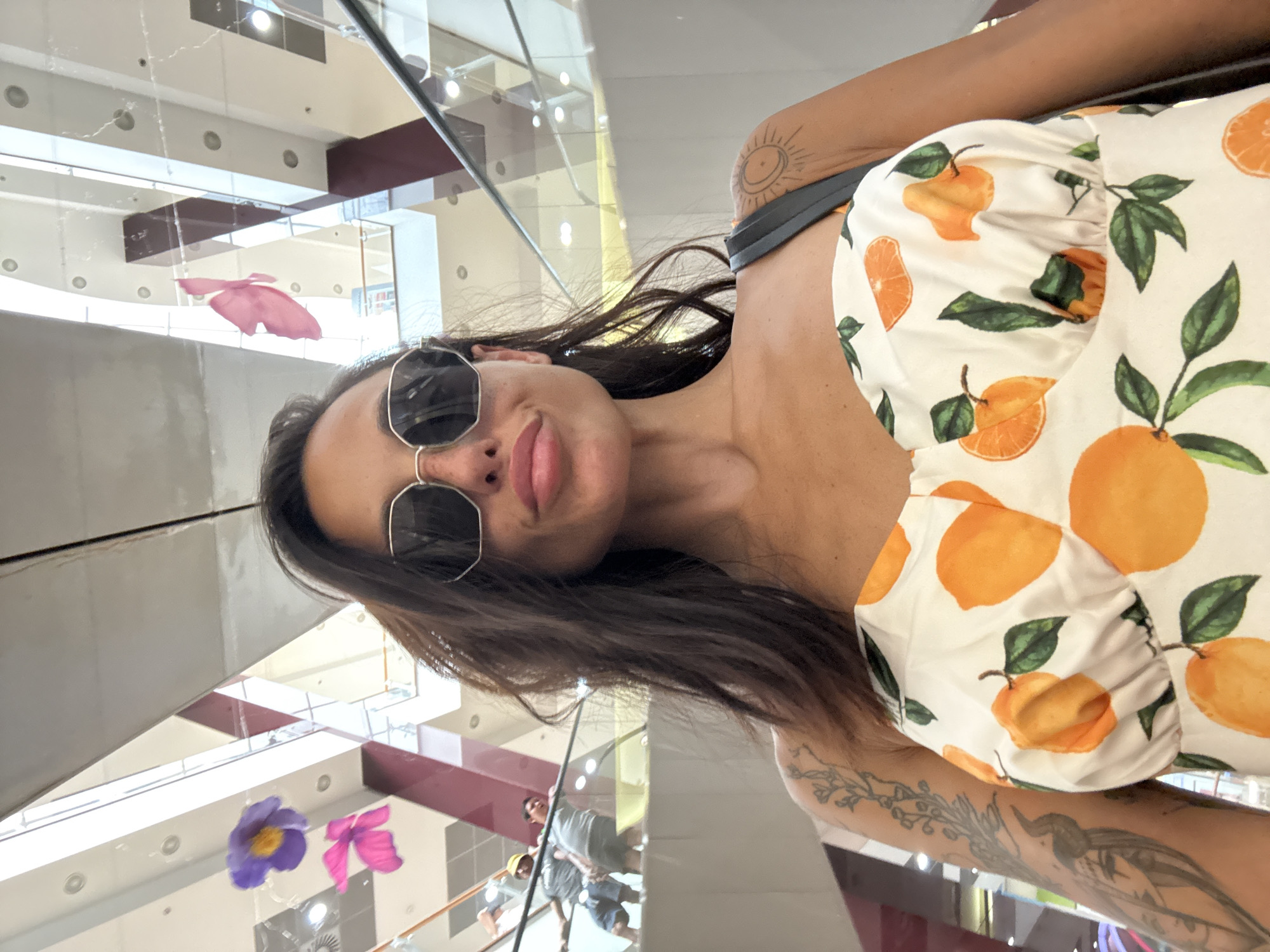A woman with long, dark hair and sunglasses takes a selfie in a well-lit, indoor location. She is wearing a white dress with an orange fruit print. She has tattoos on her arm and shoulder, including a sun design on her shoulder. The background shows a multi-level interior with escalators and railings, suggesting a shopping mall or similar public space. In the distance, two people can be seen on an escalator. Hanging from the ceiling are two decorative floral elements, one purple and one pink. The overall lighting suggests it is daytime. The image appears to be taken in Pleven, Bulgaria.