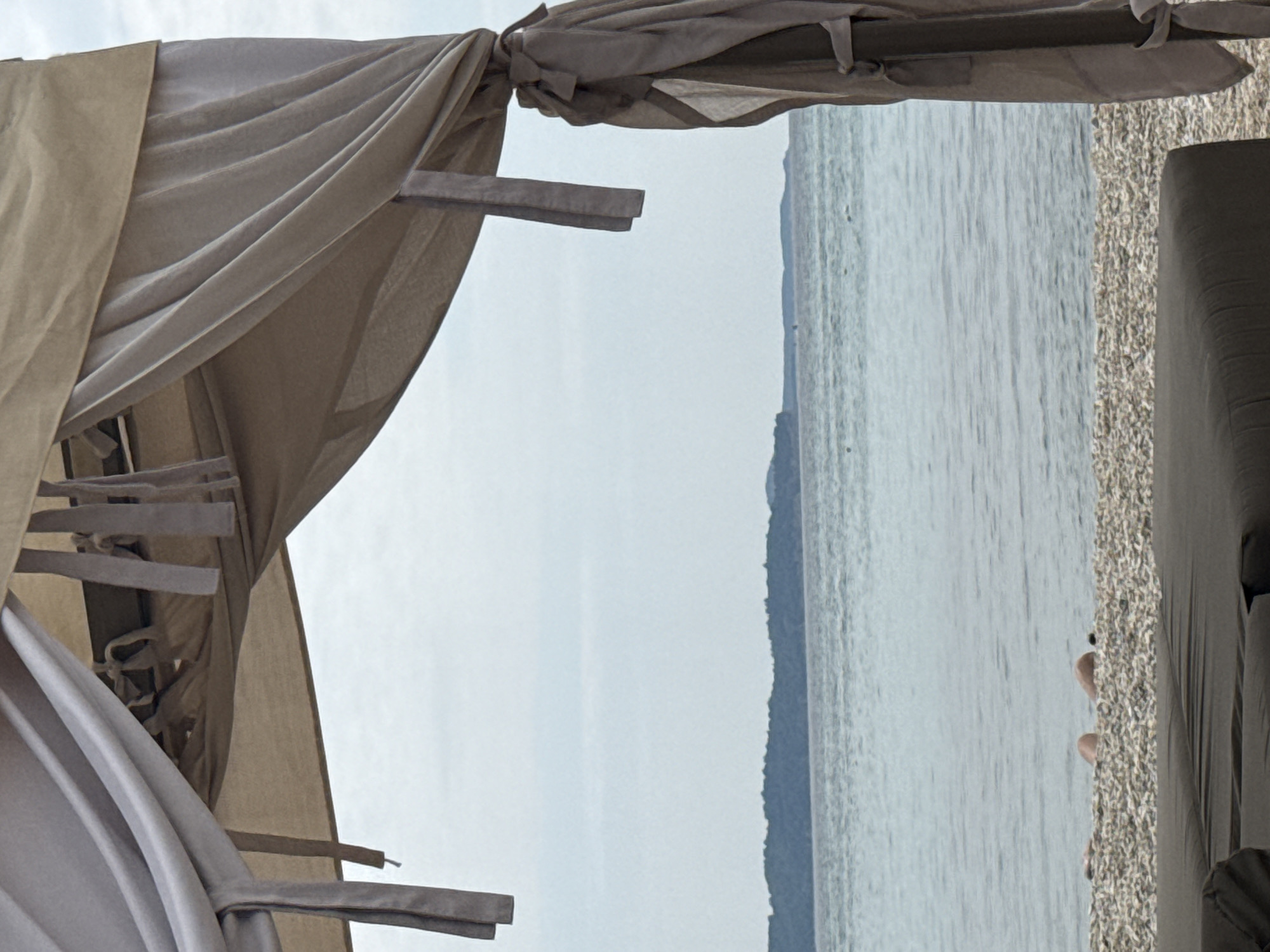 A canopy with beige fabric drapes over a beach in Limenaria, Greece. The canopy casts shadows on the pebbled beach, where the legs of a person are visible. The sea stretches out to the horizon, meeting a blue sky. A distant, tree-covered island is visible in the background. The overall scene is serene and evokes a sense of relaxation.