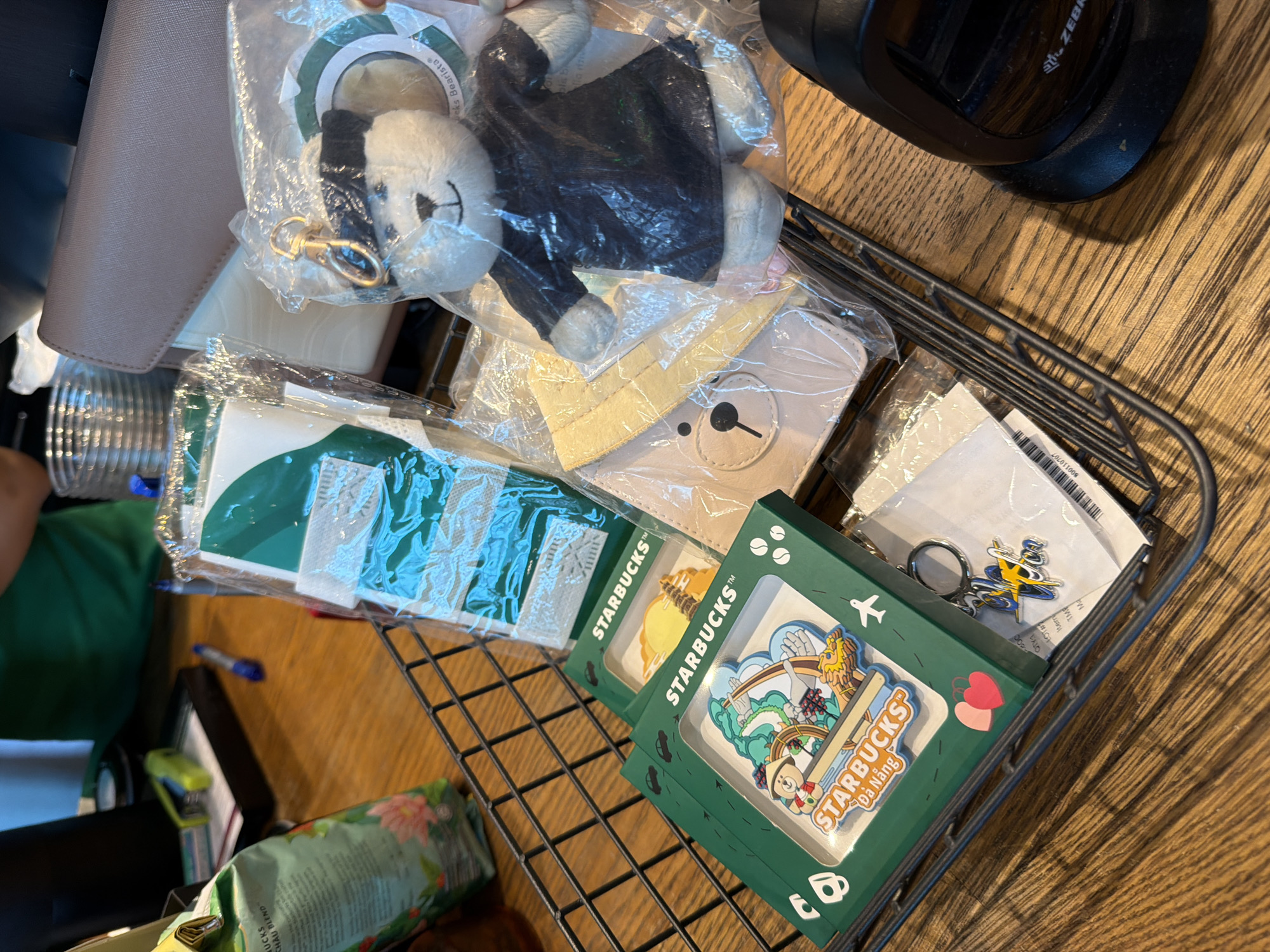A cluttered basket containing an assortment of Starbucks merchandise is positioned on a wooden surface. The basket contains a stuffed bear keychain, two green boxes of Starbucks Da Nang pins, green Starbucks napkins, a barcode scanner, Starbucks Chau Blend coffee bag, a stapler, pens, and miscellaneous items. The Starbucks Da Nang boxes feature images of a bear in various settings. The scene is in Nha Trang Ward, Vietnam.