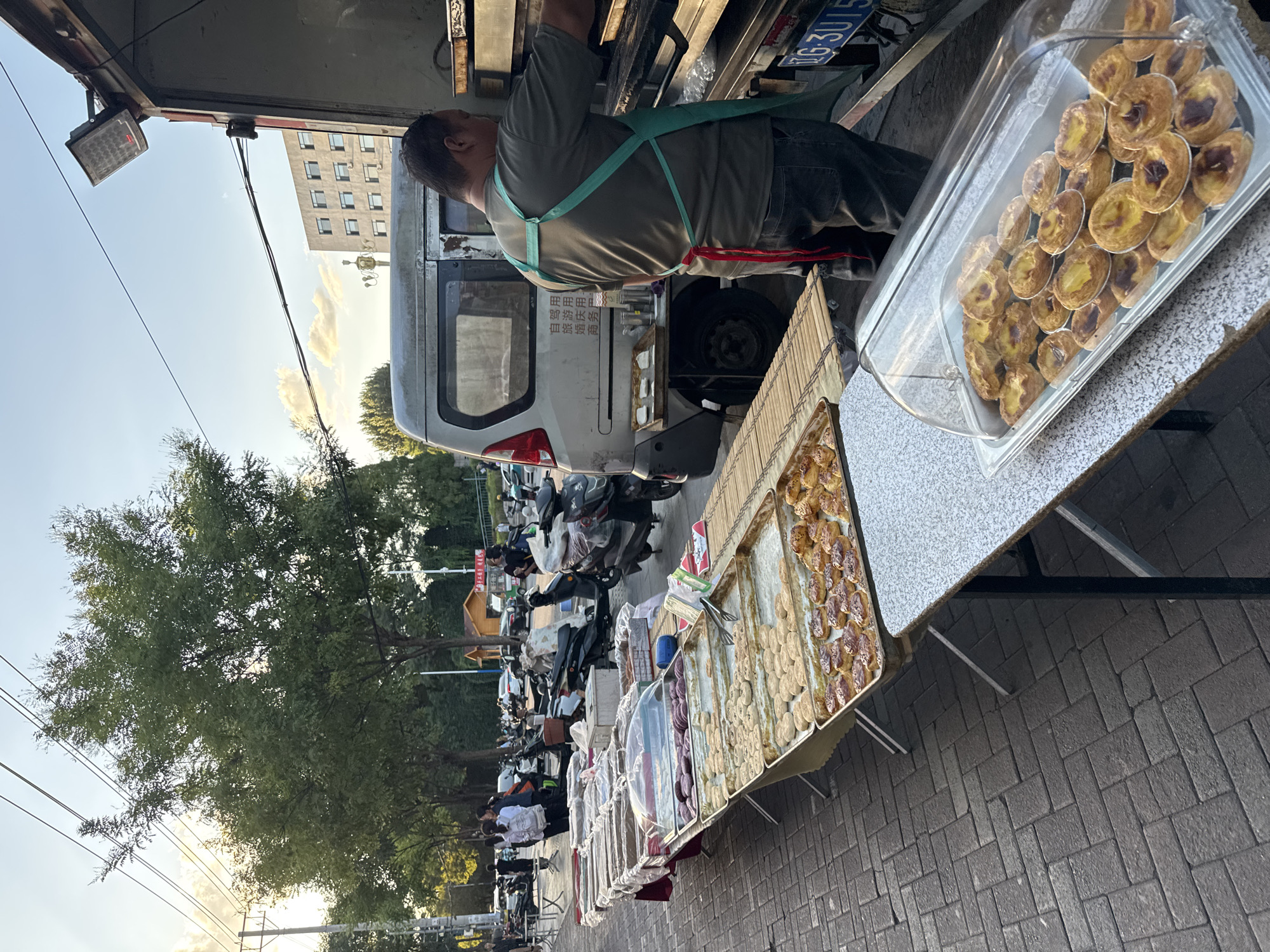 This image depicts an outdoor food stall on a paved sidewalk in Zhongtun, China. A man wearing an apron is standing behind a food truck, possibly preparing or serving food. In front of the food truck, multiple tables are laden with various food items, primarily baked goods. These include trays of what appear to be pastries, possibly egg tarts, and other round, light-colored baked goods. A variety of dishes and containers are also visible on the tables, covered with plastic wrap and lids. In the background, trees line the street, and several motorcycles are parked. Buildings and utility poles are also present, suggesting an urban or semi-urban environment. The lighting indicates it may be late afternoon or early evening.