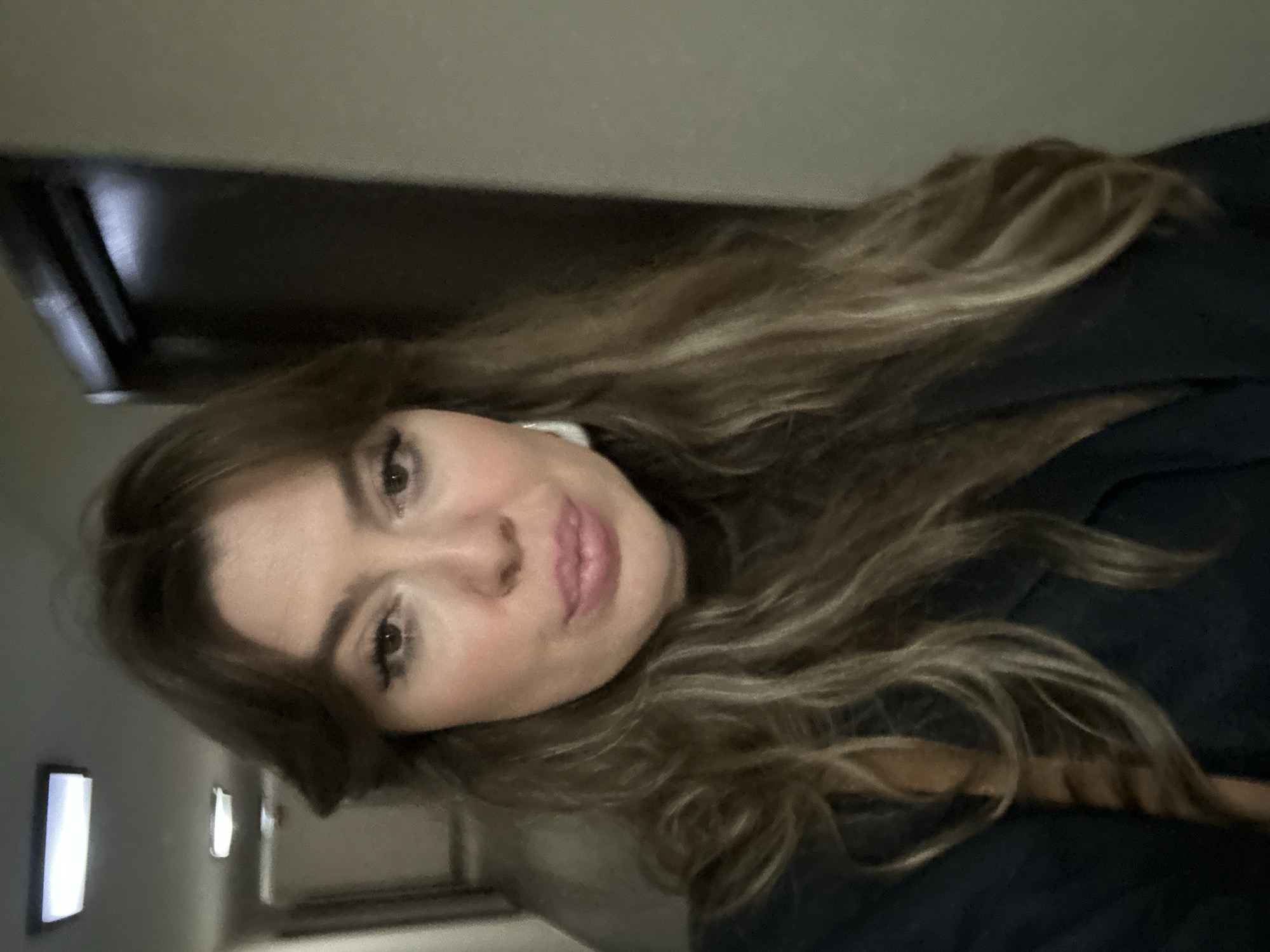 A woman with long brown and blonde hair is looking at the camera. She is wearing a dark-colored jacket. The background appears to be a hallway with a door on the left. It is not possible to determine the location in Batumi, Georgia from this image.