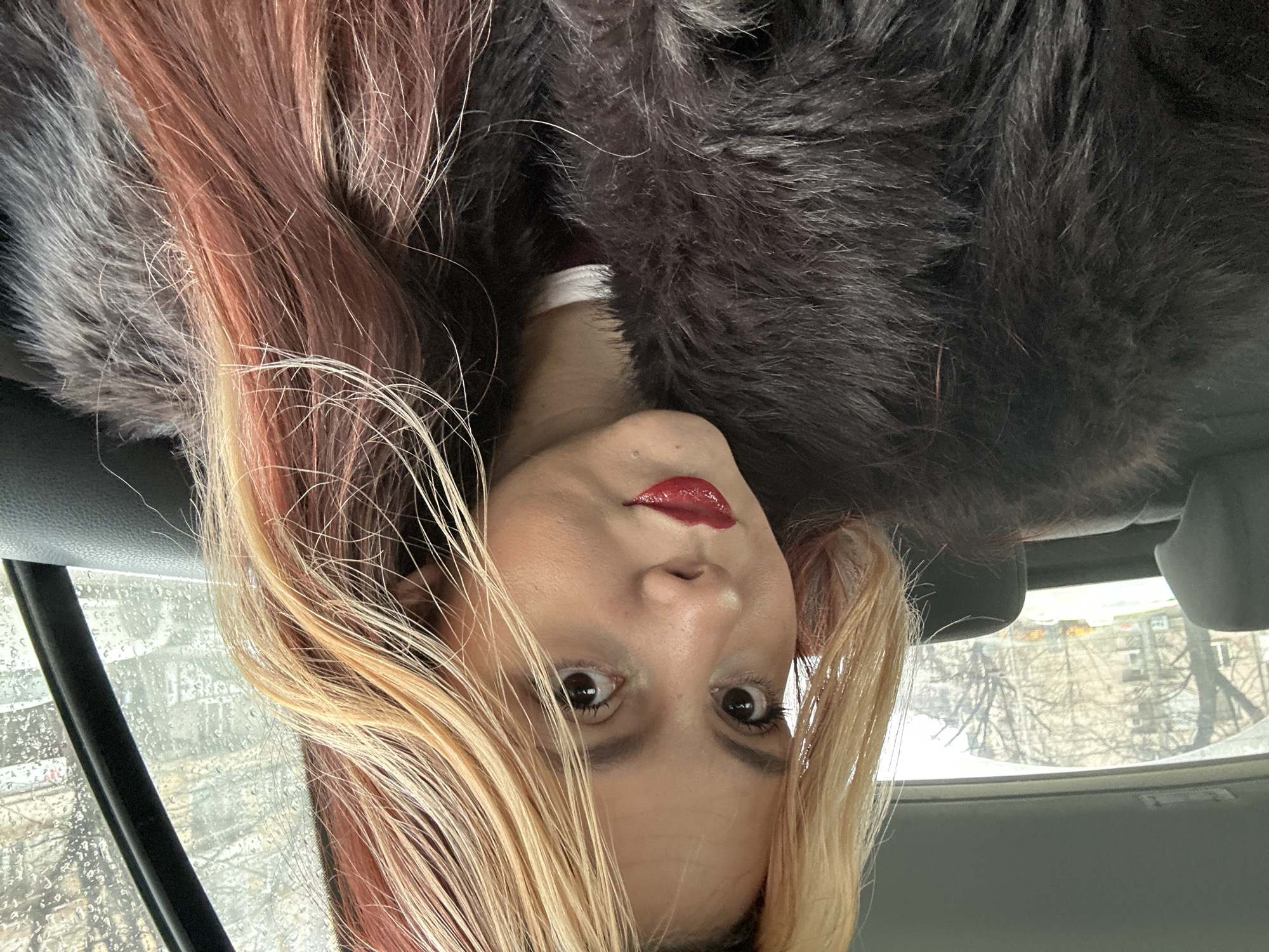 A female with blonde and red-streaked hair is shown in a close-up, upside-down selfie. She is wearing red lipstick and a dark, fur-like garment. The background appears to be inside a car, with a rain-streaked window and bare trees visible outside. Based on the architecture and the type of trees, the location could be in Sofia, Bulgaria.