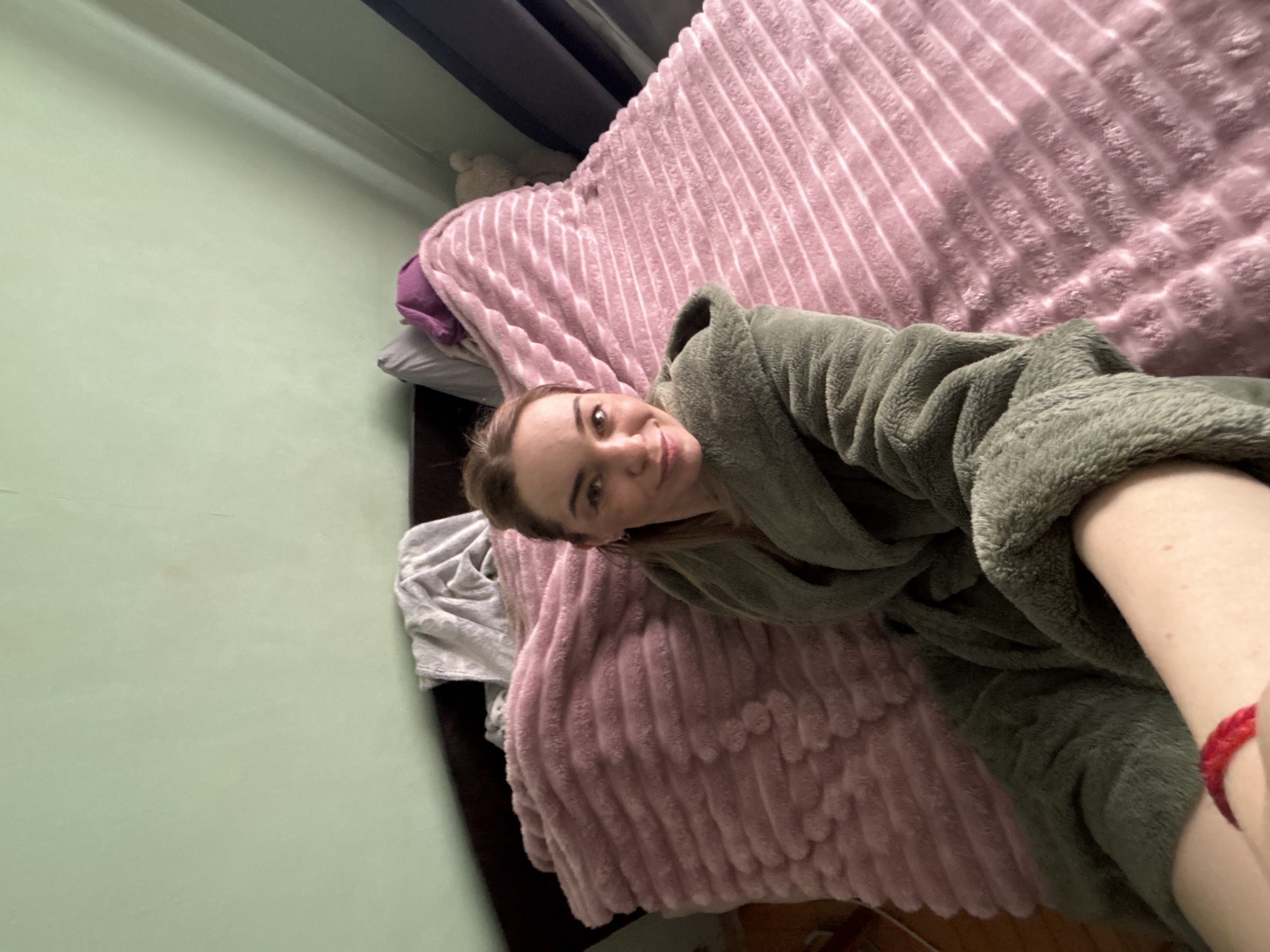 A person is lying on a bed with a pink, textured blanket. The person is wearing a green robe. A portion of their leg and red wristband are visible. The background includes a light green wall and part of a dark-colored bed frame. A stuffed toy is visible on the top edge of the headboard. The location is likely indoors. Based on the provided information there is no indication this photo was taken in Vsevolozhsk, Russia.