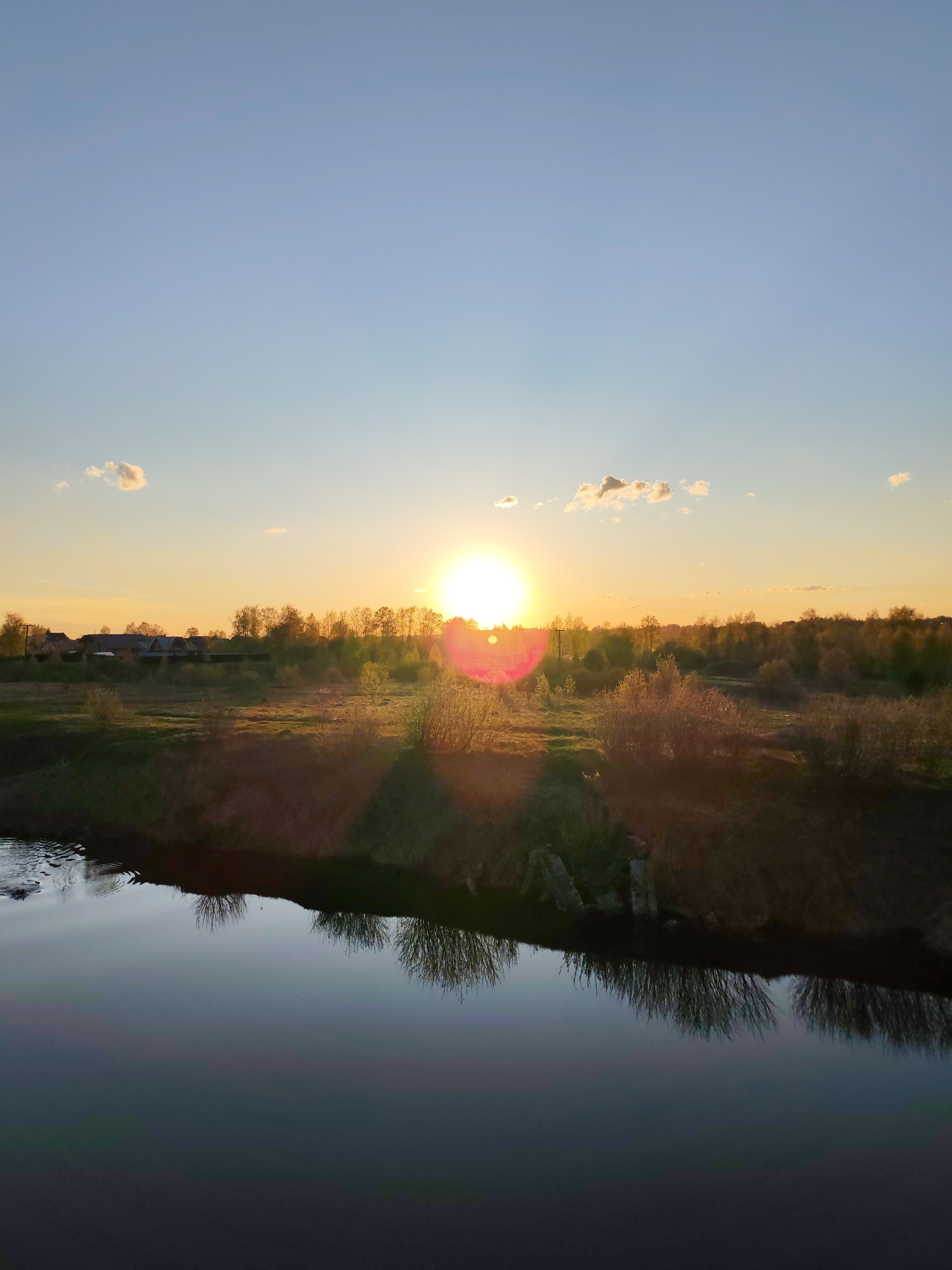 The image shows a rural landscape at sunset, possibly in Myshkin, Russia. A calm body of water, likely a river or canal, reflects the vegetation along its banks. The opposite bank is covered in trees and shrubbery, with hints of buildings or structures in the distance. The sun is setting, creating a bright glare and a warm glow over the scene, with some clouds in the upper portion of the sky. The foreground is out of focus.