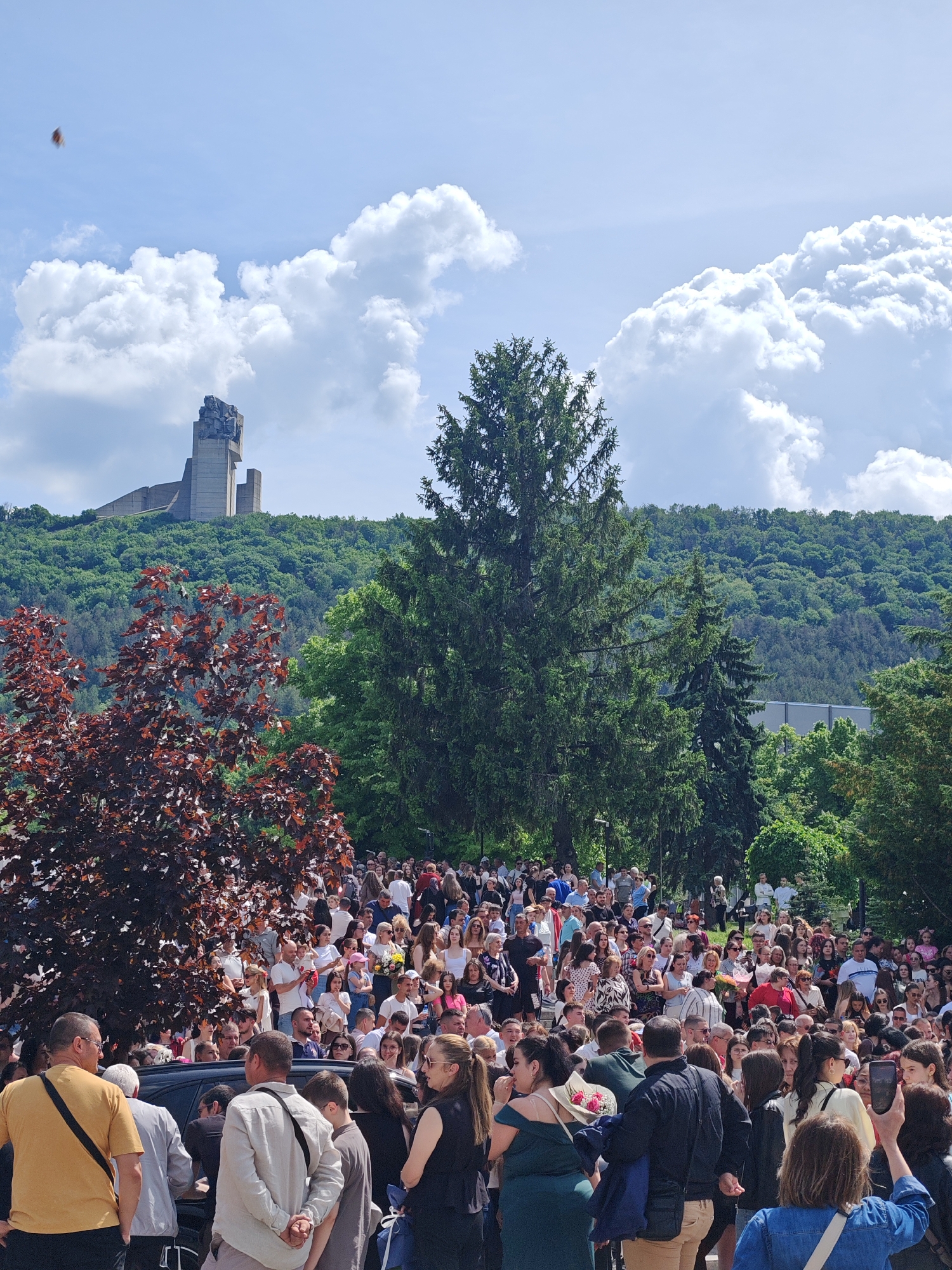 The image captures a large crowd of people gathered in an outdoor setting, likely a park or public space in Shumen, Bulgaria. In the background, the Monument to 1300 Years of Bulgaria on the Shumen Plateau is visible. The crowd is predominantly composed of young people, likely students or recent graduates, along with some older individuals. Many are dressed in formal or semi-formal attire. Some individuals are holding flowers, suggesting a celebratory event, possibly a graduation or commencement ceremony. People are standing, talking, and interacting with each other. In the foreground, a few individuals are partially obscured. A dark-colored vehicle is also visible near the front of the crowd. The weather appears sunny, with a blue sky and puffy white clouds. Trees surround the crowd, and the terrain slopes upward towards the monument.