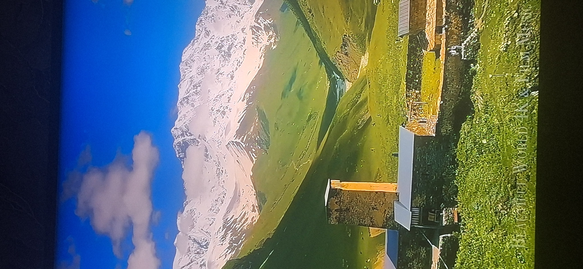 The image depicts a mountainous landscape with a village in the foreground. A tall, stone tower stands prominently in the village, along with several stone houses with metal roofs. The mountains in the background are snow-capped and rugged, with green valleys in between. A river or stream is visible flowing through one of the valleys. The sky is a bright blue with scattered clouds. There is text at the bottom of the image that reads "BLACKHAT WORLD 4K & FILM MES". The image is not located in the city of Bila Tserkva, Ukraine.