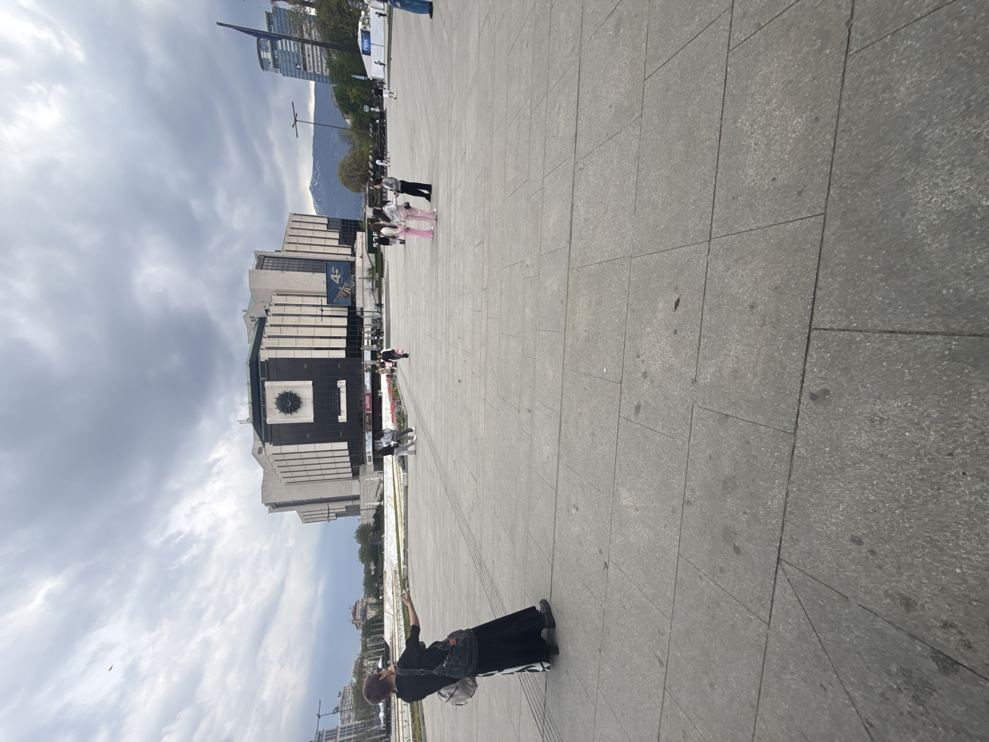 An outdoor wide shot, rotated 90 degrees clockwise, captures a large paved plaza in Sofia, Bulgaria, under an overcast sky. In the foreground, a person with dark clothing and a backpack stands, holding a phone up as if taking a picture or video. Their back is to the viewer, and they are facing a large, modern building.
This prominent building, located to the left, features a striking black and white facade with architectural details, including a large circular emblem on its upper black section. This is identifiable as the National Palace of Culture (NDK) in Sofia. A banner on the building displays what appears to be "NPMC" or "NDK" and "4G". Another banner to the far left has the text "NRSC".
Further into the mid-ground, two individuals, one in a grey top and black trousers, and another in a pink tracksuit, are walking across the plaza. Other people are scattered further in the background. The expansive grey paving of the plaza occupies a significant portion of the frame, showing distinct tile patterns and some minor stains.
In the distance, behind the buildings, large mountains (Vitosha Mountain) are visible, partially obscured by the cloudy sky. Some smaller trees and shrubs are also present near the base of the mountains and around the plaza. A light pole stands in the middle distance. The overall impression is a bustling public space on a cool, cloudy day. The word "SOFIA" is also visible on a small sign or structure in the mid-ground.