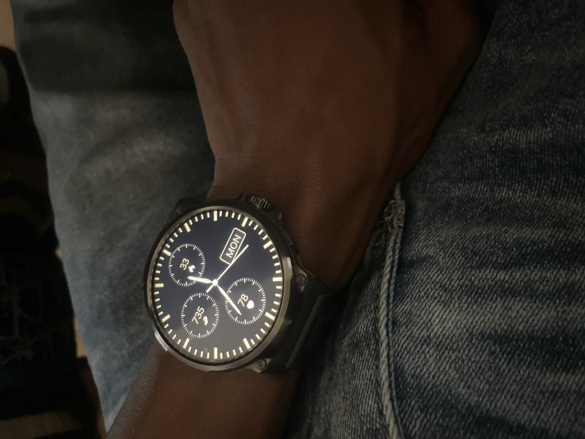 The image displays a close-up of a person's wrist wearing a smartwatch. The smartwatch has a dark face with white markings and indicators. Prominently visible are the numbers "735", "33", and "78", along with the word "MON" (likely indicating Monday). The watch is worn on a wrist with dark skin, and the background consists of blue denim fabric. The scene is captured from a high-angle perspective, focusing on the watch and wrist. No specific location within Ihiagwa, Nigeria, can be identified from the visual information provided.