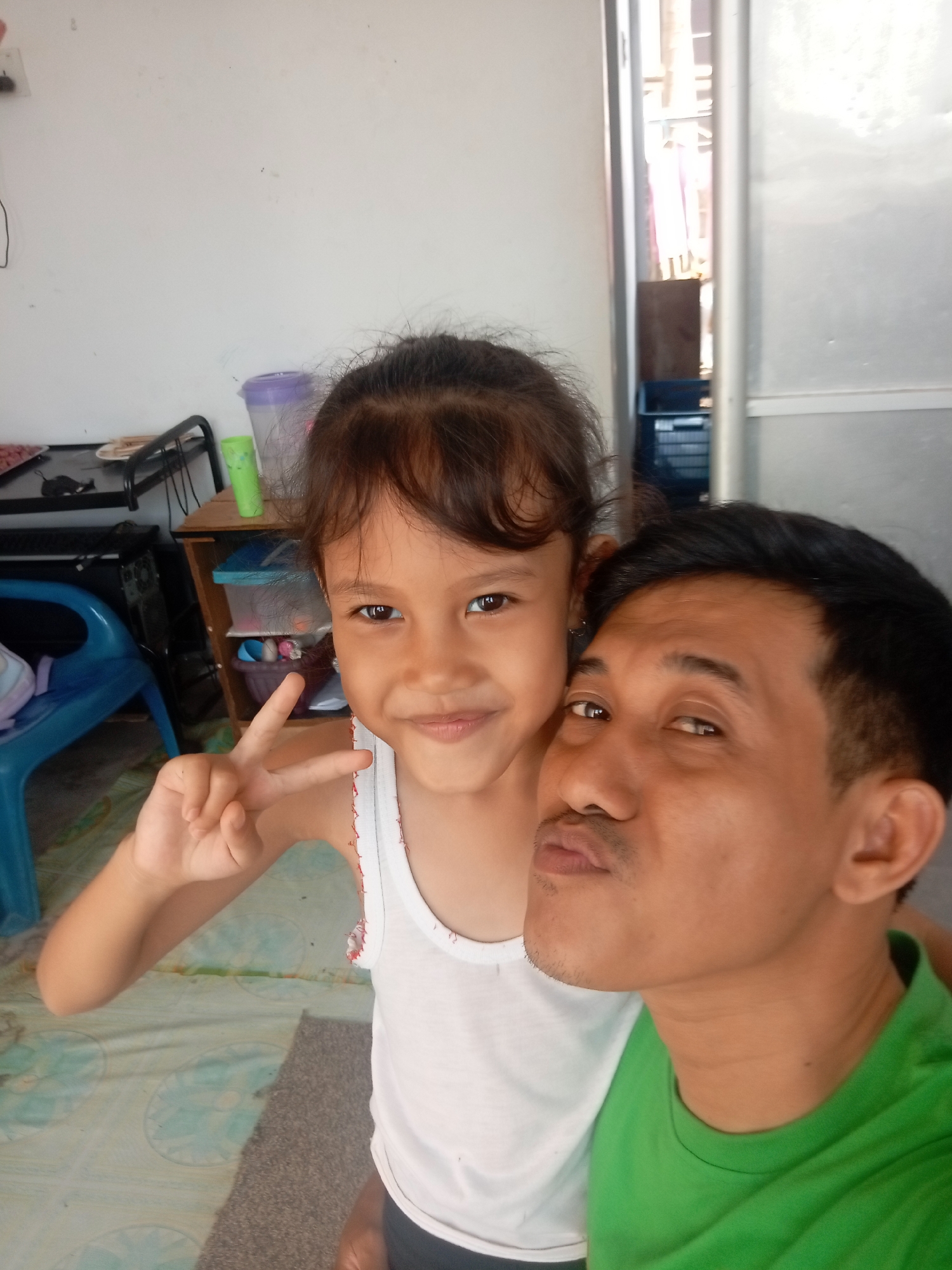 The image shows a young girl and a man posing for a selfie. The girl is in the foreground, on the left side of the image, and is wearing a white tank top. She has dark hair and is making a peace sign with her left hand. She is smiling and looking at the camera. The man is on the right side of the image, leaning in towards the camera with his mouth slightly pursed. He is wearing a green t-shirt and has dark hair. The background is blurred but appears to be an indoor setting, possibly a room in a house. There is some furniture and shelves visible, with containers and other items on them. The lighting suggests it is daytime. The overall mood of the image is casual and friendly, with both subjects appearing happy. There is no visible text in the image. The location context provided is Batam, Indonesia.