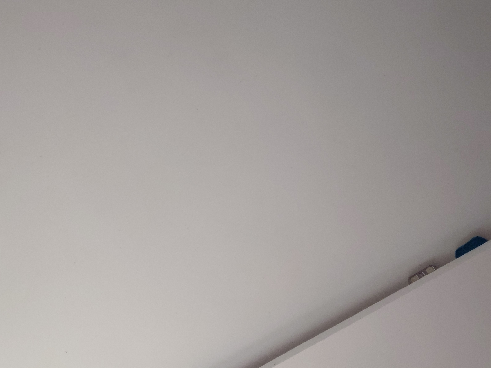 The image presents a minimalist, static view predominantly filled by a large, smooth, light-grey or off-white surface, which appears to be an interior wall or ceiling. Subtle textures and minor specks are visible across this expansive surface. In the bottom right corner, a sharp diagonal line cuts across the frame, delineating the main surface from a lower, slightly brighter white plane, likely the top edge of a piece of furniture such as a cabinet or shelf.
Resting on this lower surface, partially obscured by the diagonal edge, are two small objects. To the left, a metallic item with multiple small, rectangular segments is visible, possibly a clip, hinge, or part of a fixture. Further to the right, a small, distinct piece of a blue object, potentially made of plastic or fabric, is noticeable.
The lighting in the image is even and bright, suggesting a well-lit indoor environment, either during daytime or under ample artificial illumination. There are no people, specific activities, or discernible emotions conveyed. No text is visible within the frame. While the image itself offers no specific visual cues regarding its geographic location, it is identified contextually as Santa Ana, Venezuela.