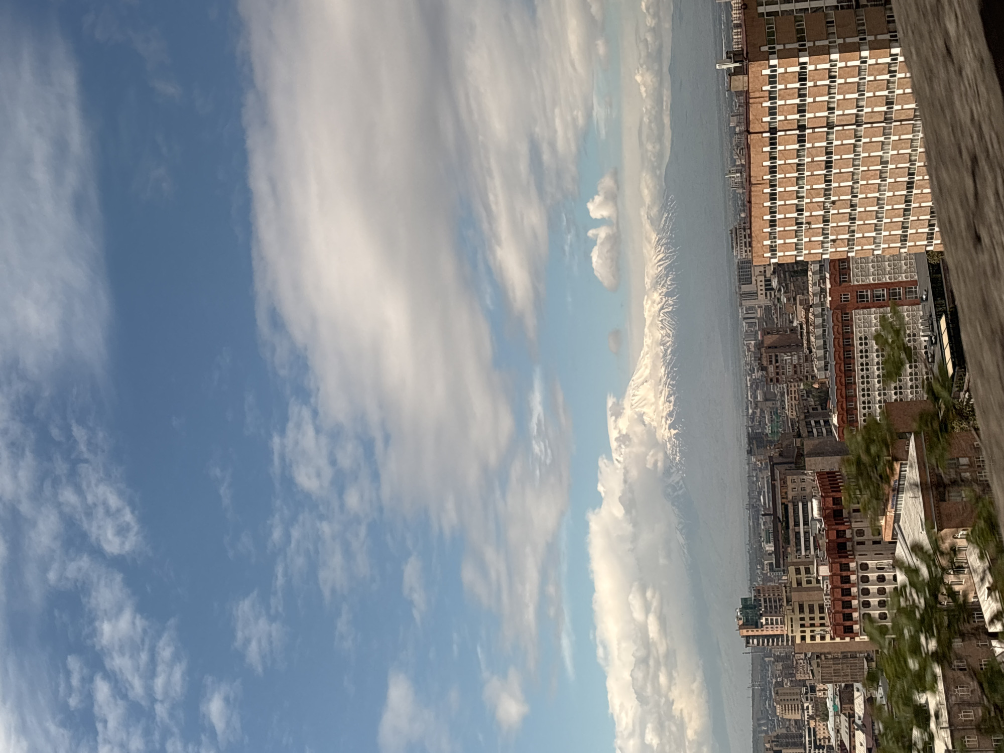The image shows a cityscape in Yerevan, Armenia, with a view of Mount Ararat in the distance. The city buildings vary in height and color, including brick and stone structures. The sky is blue with streaks of white clouds. Tall buildings stand in the foreground, partially obscuring the cityscape. Some greenery from trees is visible.