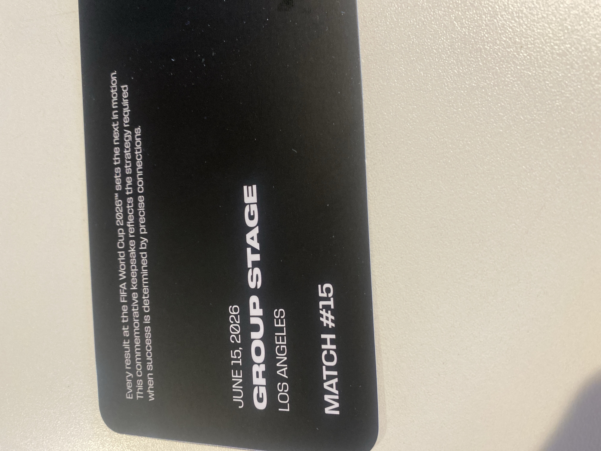 The media file shows a black ticket stub, likely a souvenir or ticket for a sporting event. The text on the ticket indicates it is for the FIFA World Cup 2026, specifically a "GROUP STAGE" match. The date is June 15, 2026, and the location is Los Angeles, United States of America. The ticket also specifies "MATCH #15." Along the left side of the ticket, there is a descriptive paragraph about the event, noting that "Every result at the FIFA World Cup 2026 sets the next in motion" and that the keepsake reflects the strategy required for success through "precise connections." The image is taken from a slightly angled perspective, with the ticket lying on a textured, light-colored surface. There are no people or specific scene details visible beyond the text on the ticket. The lighting appears to be ambient, typical of an indoor setting.