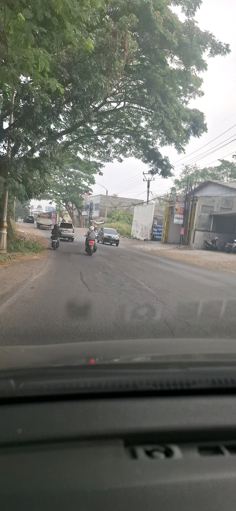 The image depicts a street scene in Rancagoong, Indonesia, viewed from the perspective of a vehicle's dashboard. The road is asphalt and shows signs of wear, with several potholes. A mature tree with lush green foliage dominates the left side of the frame, casting shadows on the road.
Several vehicles are present on the road. In the foreground, two motorcycles are moving away from the viewer. The motorcycle on the right has a rider wearing a helmet and a grey jacket. The motorcycle on the left has a rider with a dark jacket. Further ahead, a silver SUV is traveling in the same direction. Behind the SUV, a light-colored truck is also on the road. On the right side of the road, a dark-colored sedan is visible.
Buildings line the right side of the road. One building appears to be a workshop or industrial facility with a corrugated metal roof. Another building is a lighter color with several windows. A large white wall with graffiti stands between these structures. Several parked motorcycles are visible in front of one of the buildings. Electrical poles and overhead wires are also present. The sky is overcast.