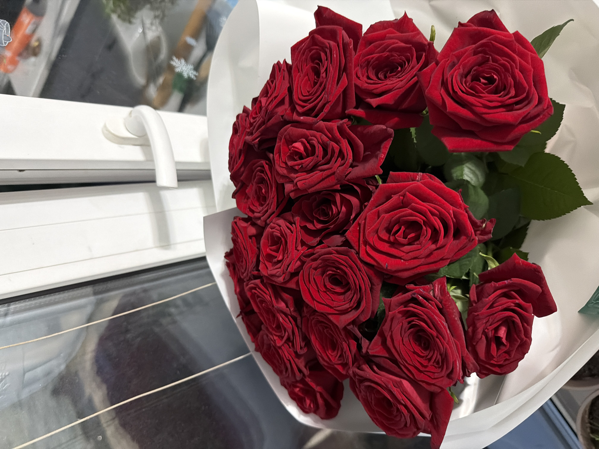 A bouquet of red roses is presented against a backdrop of a window and a white windowsill with a white handle. The roses are deep red with velvety petals, arranged in a lush, full bouquet. Some roses are fully bloomed, showcasing intricate layers of petals, while others are in the process of opening. The bouquet is wrapped in white paper, which contrasts with the vibrant red of the flowers. The window behind the bouquet is reflective, showing some indistinct shapes and colors from the outside, possibly indicating an urban setting. The scene is indoors, with natural light filtering through the window. The location is specified as Tujmazy, Russia.