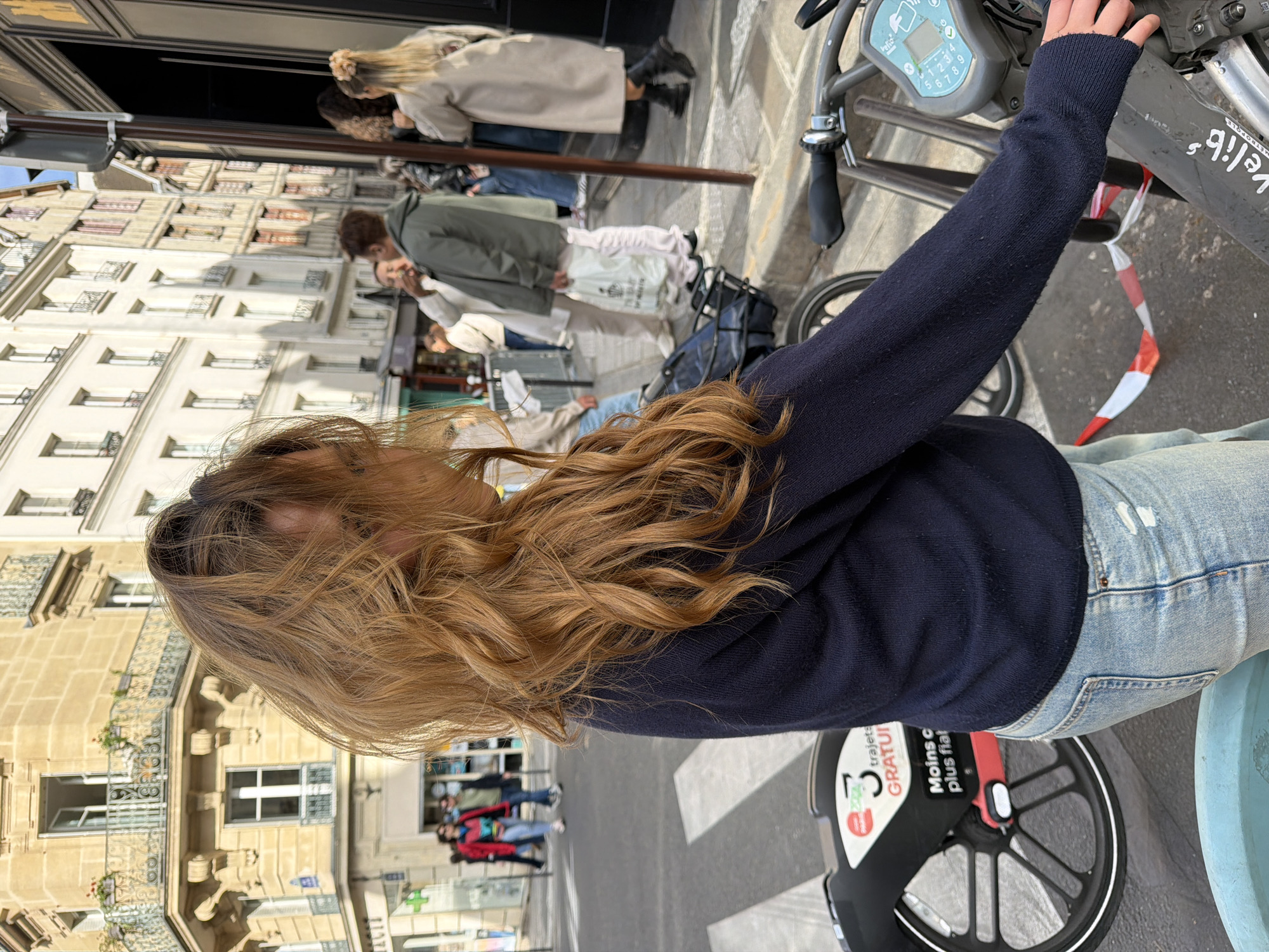 A woman stands on a street next to a Vélib' bike sharing station in Paris, France. She has long, wavy brown hair, is wearing a dark blue sweater and light blue jeans. She is reaching out to the bike's handlebars. The Vélib' station features a gray bike with a black basket, an electronic payment console, and a front wheel with a marketing sticker. The street has a crosswalk visible in the background. Pedestrians are walking along the sidewalk and crossing the street. Parisian architecture is visible on the buildings.