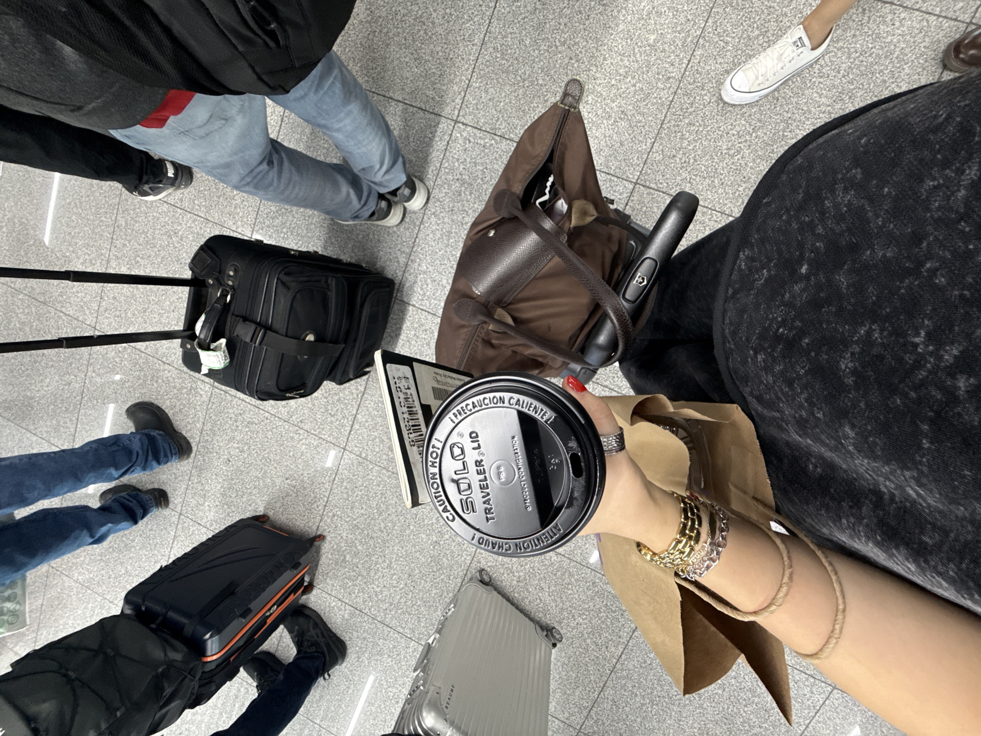 The image depicts a high-angle shot of an airport concourse. Several people with luggage are seen from the waist down, indicating they are travelers. A person is holding a coffee cup labeled "Solo Traveler Lid" and a brown paper bag. A boarding pass or ticket is held next to the cup. The airport floor is tiled in a grid pattern. The surroundings suggest a busy travel hub, though the specific location within Mexico City is not determinable from the visual data.