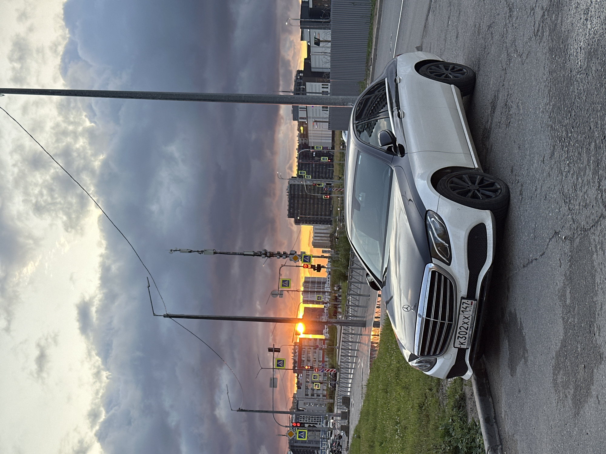 This is a vertical, eye-level, outdoor photograph taken in Sinyavino, Russia, during sunset. The main subject is a white Mercedes-Benz E-Class sedan parked on the side of an asphalt road. The car is facing away from the camera, with its front right quarter visible. It has black wheels and a black hood. The license plate, which is partially visible, reads "K302XX 147".
The background features a dramatic sky filled with dark, heavy clouds, characteristic of stormy weather. However, the sunset breaks through, casting a warm, orange glow on the horizon, illuminating a city skyline with numerous apartment buildings. Traffic lights and pedestrian crossing signs are visible along the road. The scene is dominated by the contrast between the dark, moody sky and the vibrant sunset. A tall metal pole with wires stretches across the frame, adding a strong vertical element to the composition. Green grass grows along the roadside. The overall mood of the image is one of urban twilight, with a blend of natural beauty and man-made structures.