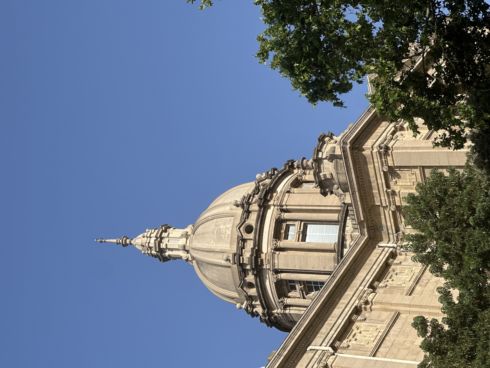 The image shows a close-up, low-angle view of the dome and upper facade of the Palau Nacional in Barcelona, Spain, against a clear blue sky. The building, made of stone, features classical architectural elements such as columns, intricate carvings, and rounded windows. A prominent dome with a finial caps the structure, and part of a large tree with green leaves is visible in the foreground, partially obscuring the building. The scene is bathed in bright sunlight, casting subtle shadows.