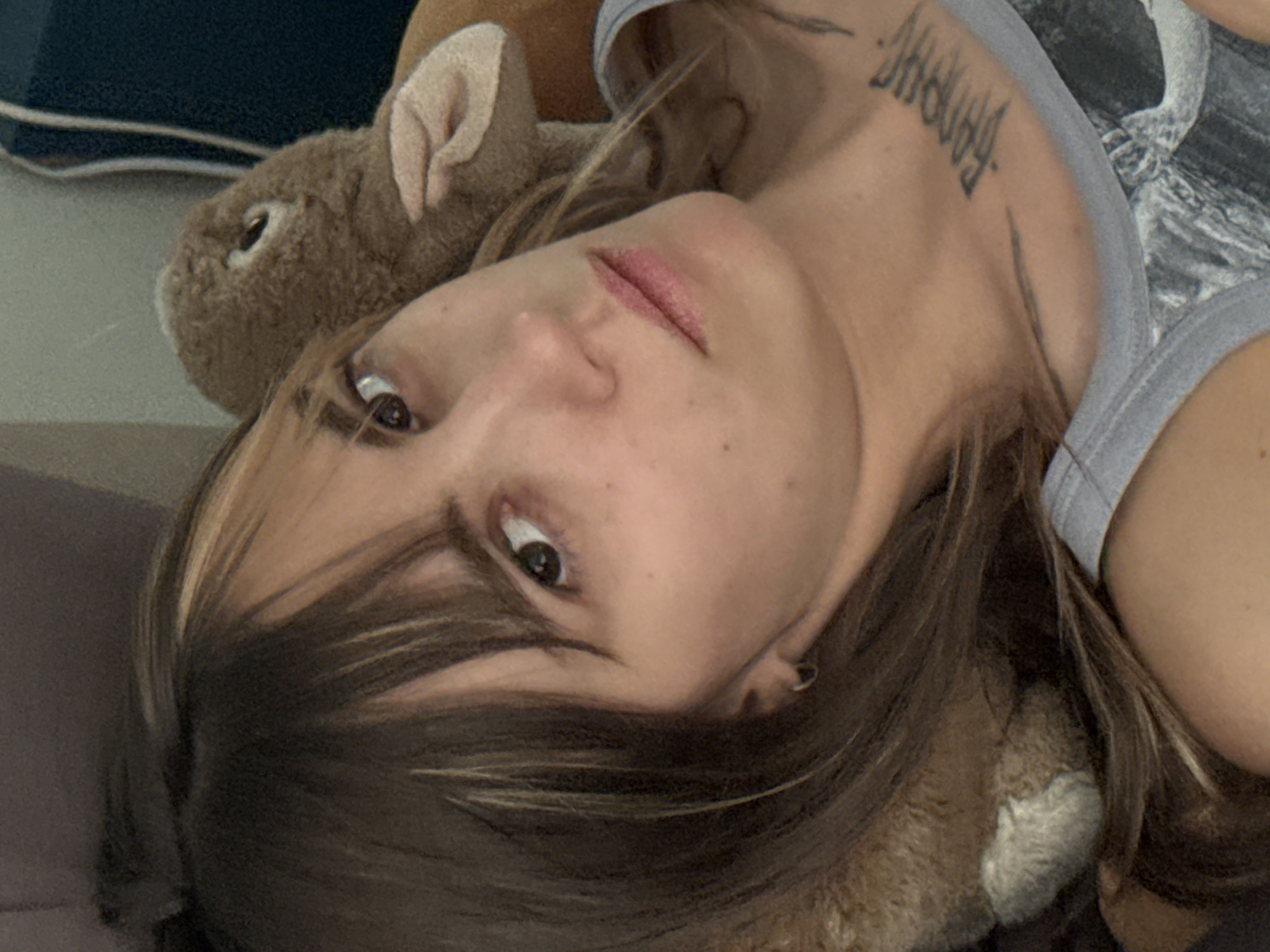 A young woman is pictured in a close-up, overhead shot. She has long, brown hair with blonde highlights and bangs. She is lying down, with a stuffed animal behind her head. She is wearing a grey tank top with a graphic print. A tattoo is visible on her collarbone. The background is blurred, but appears to be an indoor setting. There are no discernible landmarks to indicate a specific location in Moscow, Russia.