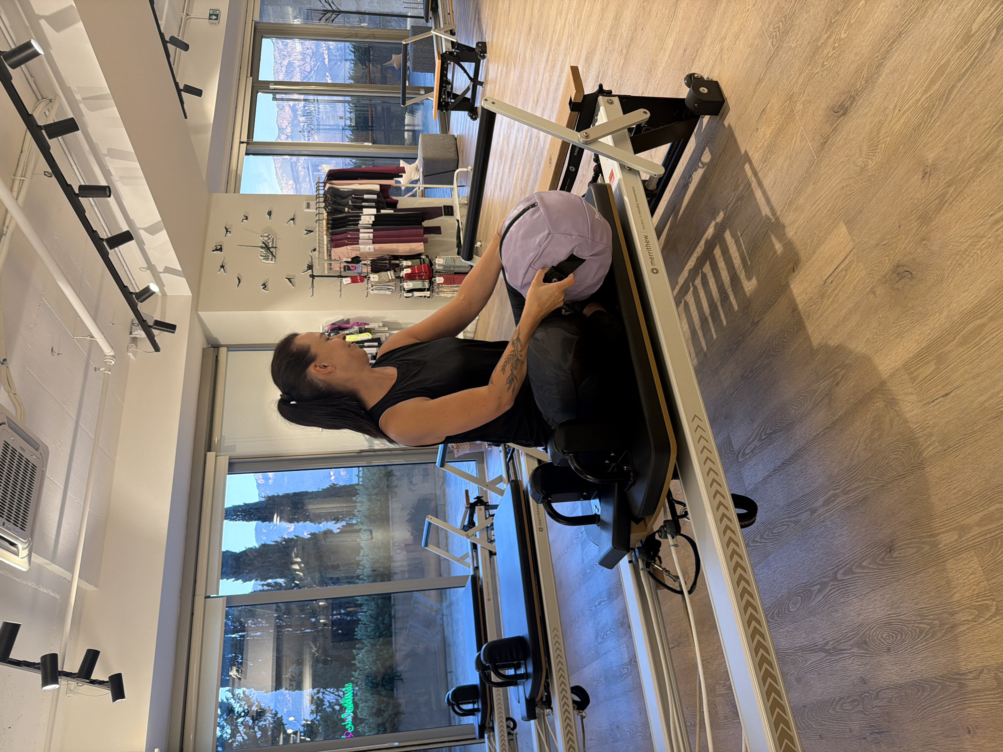 In the city of Boreti, Montenegro, a woman with dark hair tied in a ponytail is seated on a Pilates reformer. She is wearing a black tank top and grey leggings, with a tattoo visible on her left arm. She is holding a light purple weighted ball in her hands. The reformer is part of a larger Pilates studio setting, with other reformers visible in the background. The studio features large windows with a view of trees and mountainous terrain, suggesting a scenic location. The floor is made of light wood planks. The walls are white, and the ceiling has exposed lighting tracks and an air conditioning unit. Racks of athletic wear are displayed on one wall.