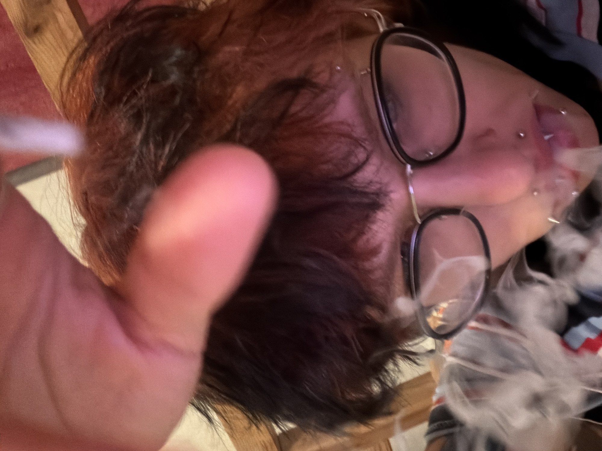 A person with dark hair and glasses is exhaling smoke. They have lip piercings. The photo is taken from a low angle with a thumb in the foreground. The background shows indeterminate items. The image is too ambiguous to determine the location in Sofia, Bulgaria.