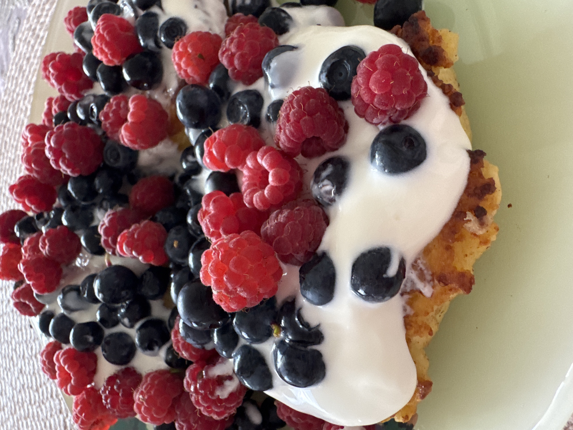 A close-up, eye-level, vertical shot shows a plate of food topped with yogurt, raspberries, and blueberries. The food appears to be some kind of pancake or fritter, with visible browned edges and a slightly fluffy interior. The white yogurt is generously spread over the food, with the red raspberries and dark blue blueberries nestled into it. The background is a textured, light-colored surface, possibly a placemat or tablecloth. A light green plate is visible beneath the food. The scene is well-lit, with natural light creating soft shadows. The focus is sharp on the food, blurring the background slightly. There are no people or animals visible in the image. The location is described as Ladozhskoe Ozero, Russia, but there is no visual information in the image that confirms this location.
