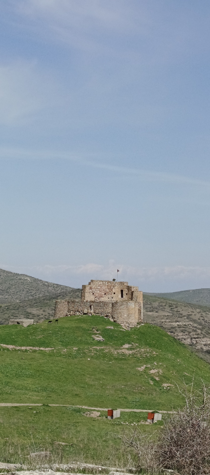 The image displays a historic stone fortress perched atop a verdant hill, under a vast, clear blue sky with minimal wispy clouds. The fortress, constructed from reddish and light brown stones, features several connected structures, including round and angular sections, suggesting its ancient or medieval origins. A small, dark flag flies from the highest point of the fortification.
The hill leading up to the fortress is covered in bright green grass, indicative of spring or early summer. Below the fortress, on the grassy slope, two dark, upright rectangular signs are visible, likely providing information to visitors. In the lower foreground, two box-like structures with reddish-orange lids and grey bases sit on the grass; these could be beehives or utility boxes. Dry, thorny bushes occupy the immediate foreground, adding a contrasting texture.
Beyond the main hill, a landscape of rolling, sparsely vegetated hills extends into the distance under a slightly hazy sky. The scene is devoid of people, presenting a tranquil view of the historical site. The time of day appears to be bright daylight with fair weather. No discernible text is visible on any objects. The setting is a rural, hilly area, consistent with the specified location of Berdavan, Armenia.