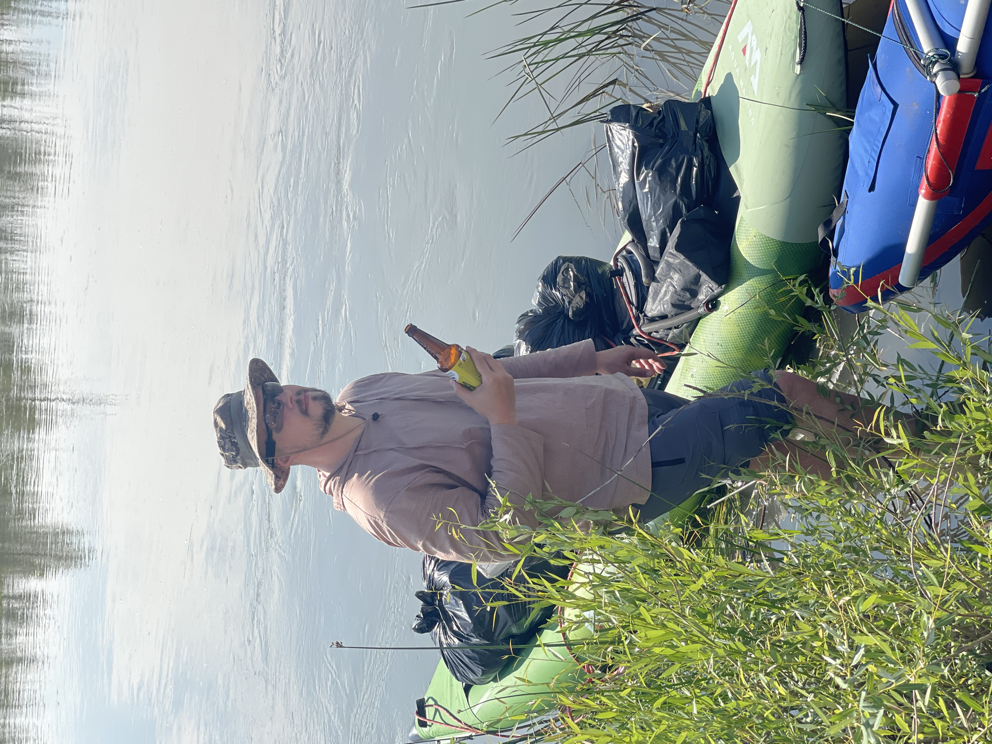 A man wearing a camouflage bucket hat, sunglasses, a tan hooded shirt, and gray shorts stands in shallow water next to a green inflatable raft. The raft is loaded with several large black trash bags. A blue inflatable raft is partially visible in the foreground, in front of the green raft. The man is holding a brown glass bottle in his right hand. He is looking to his right. The water appears calm with reflections of trees on the surface. The scene is outdoors with green vegetation on the left side of the image. The location is identified as Nezhinka, Russia.